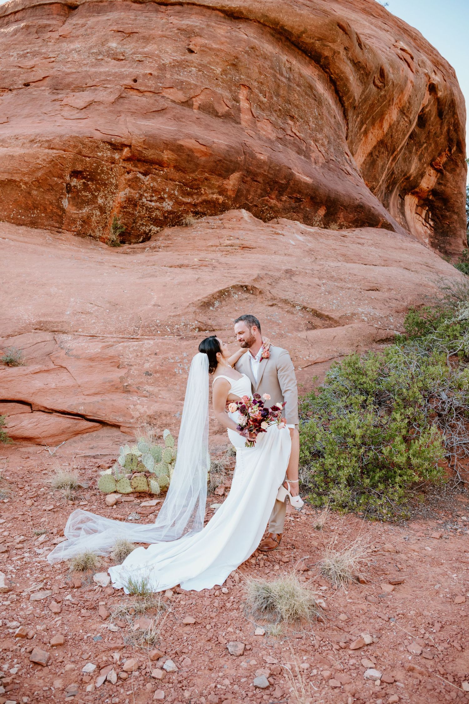 Two people in wedding attire stand together against stunning red rock formations in a desert landscape.