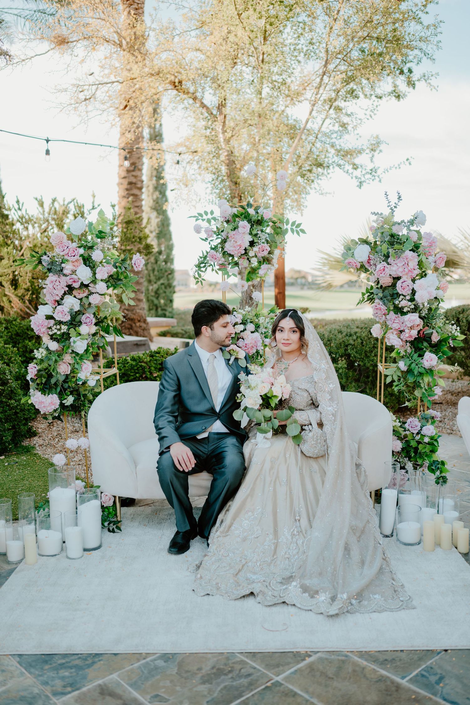 A romantic outdoor wedding scene with floral arches, white candles and a loveseat adorned with pink and purple blooms.