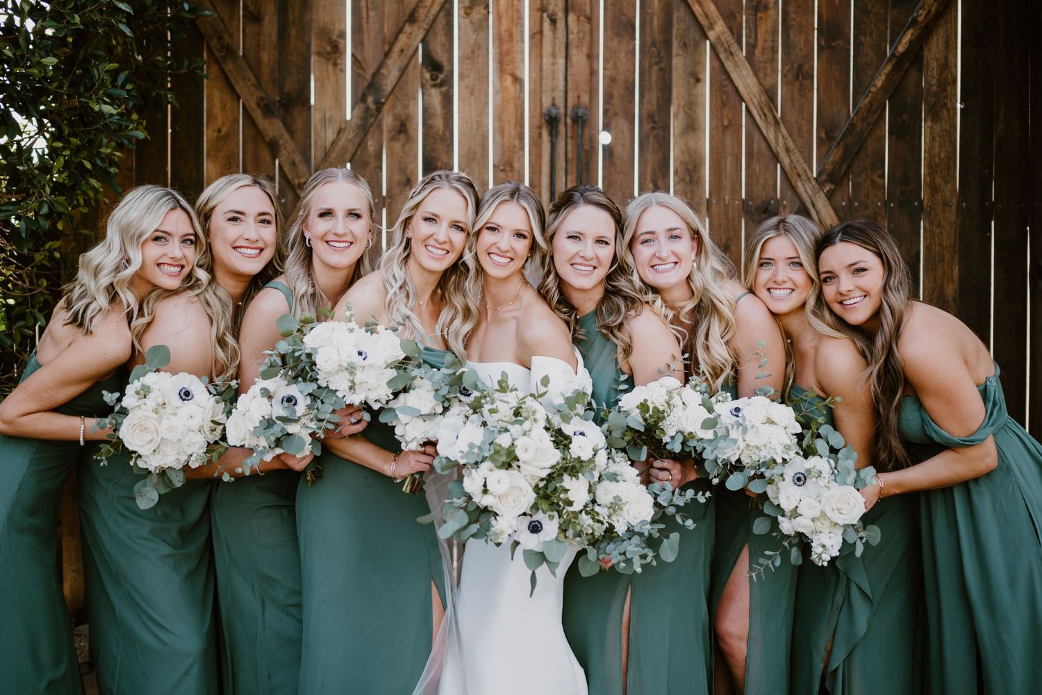Bridesmaids in matching green dresses hold white floral bouquets while posing together against rustic wooden barn wall.