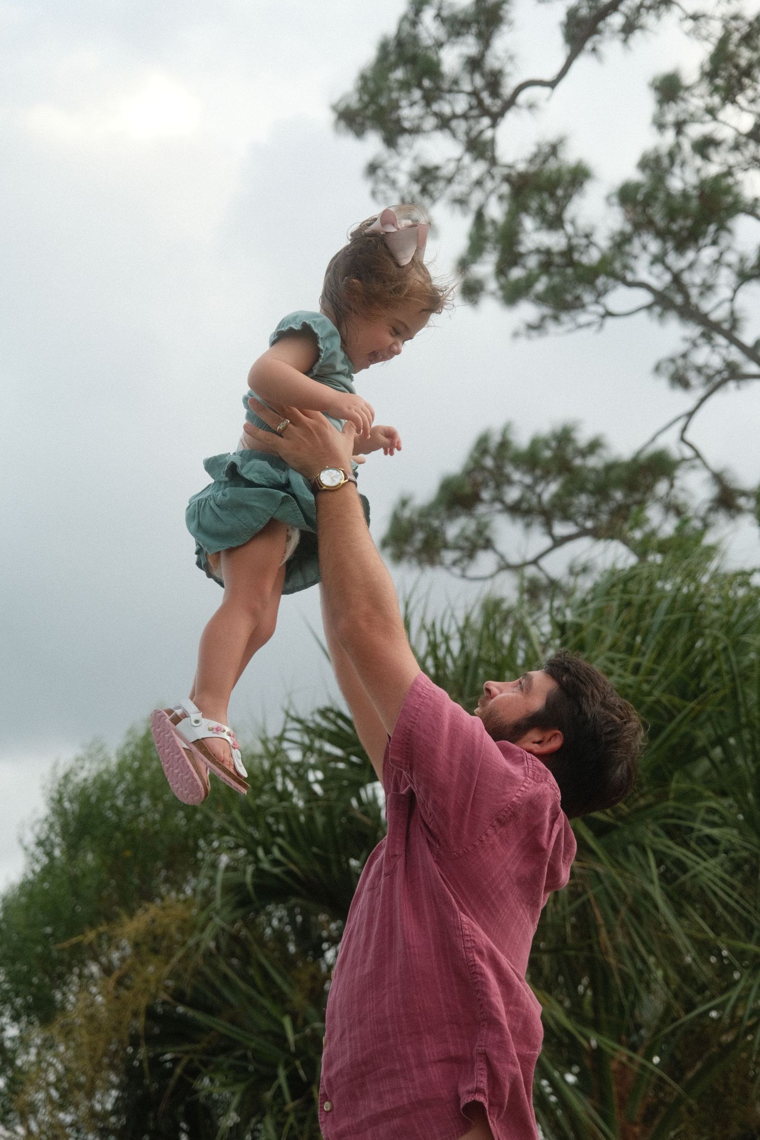 A person in a burgundy shirt lifts a small child wearing teal clothing into the air against a cloudy sky.
