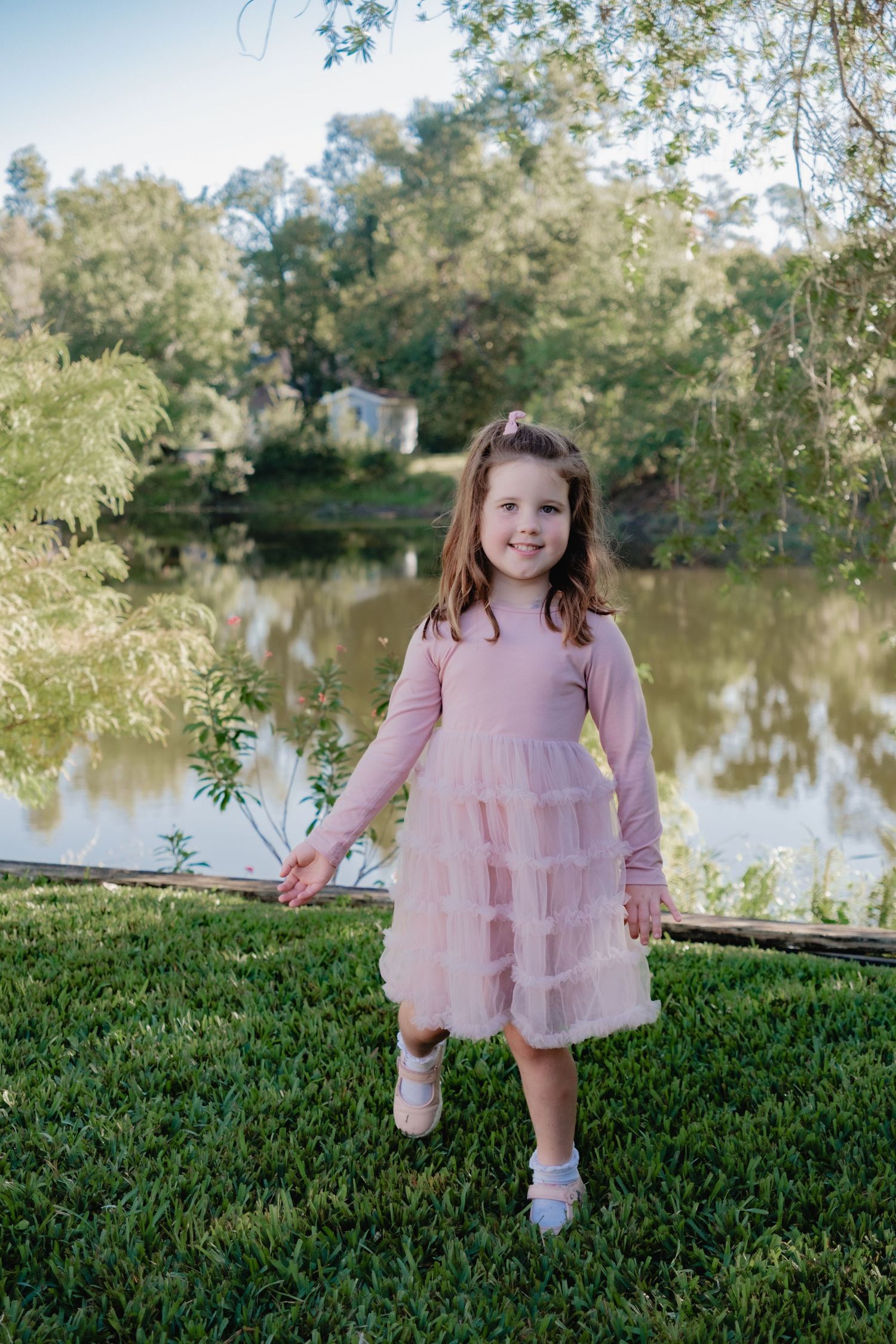 Young child in a pink tulle dress stands on grass near a tranquil pond on a sunny day.