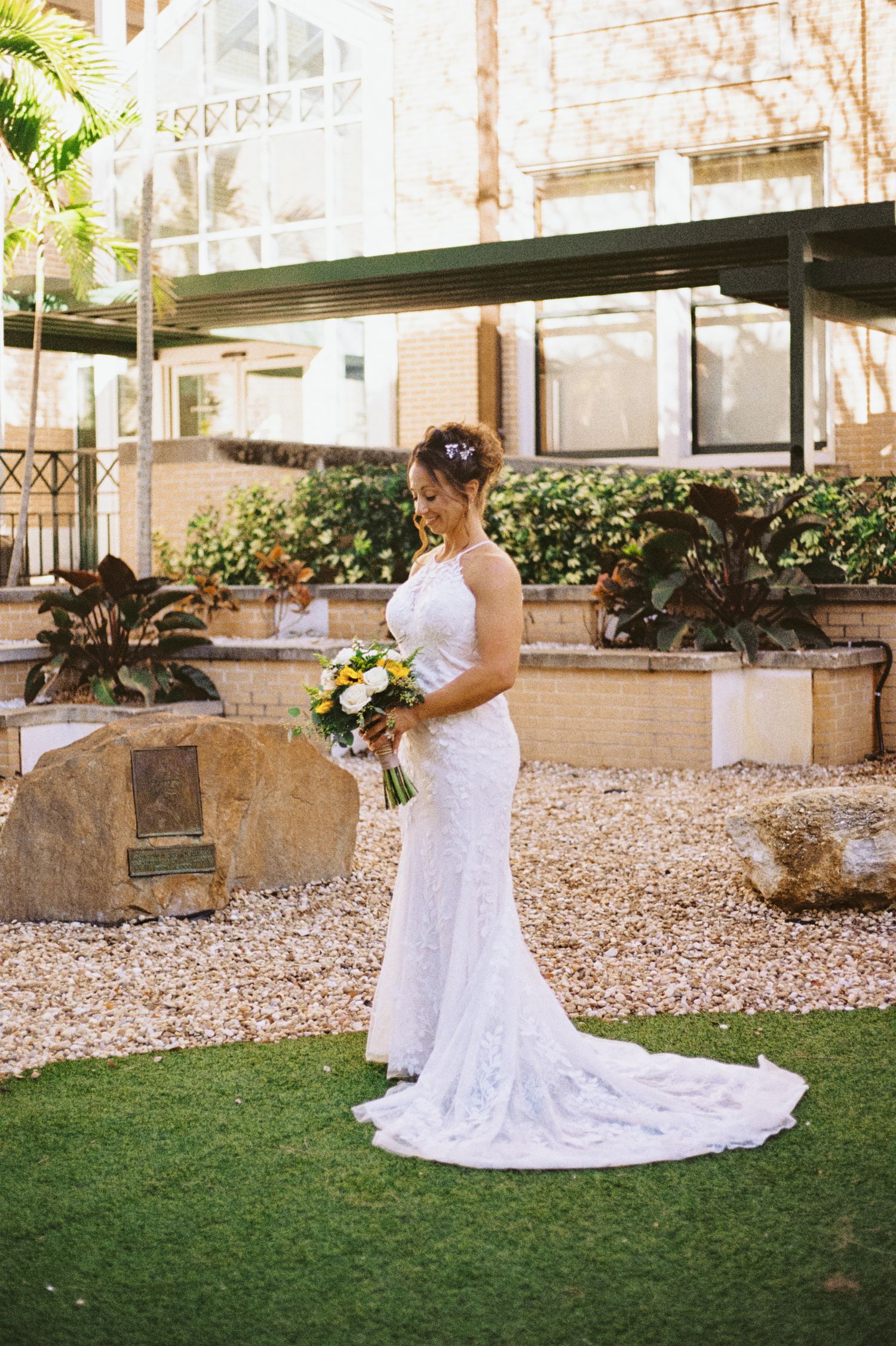 Bride in white lace mermaid gown holding colorful bouquet standing in garden courtyard on wedding day.