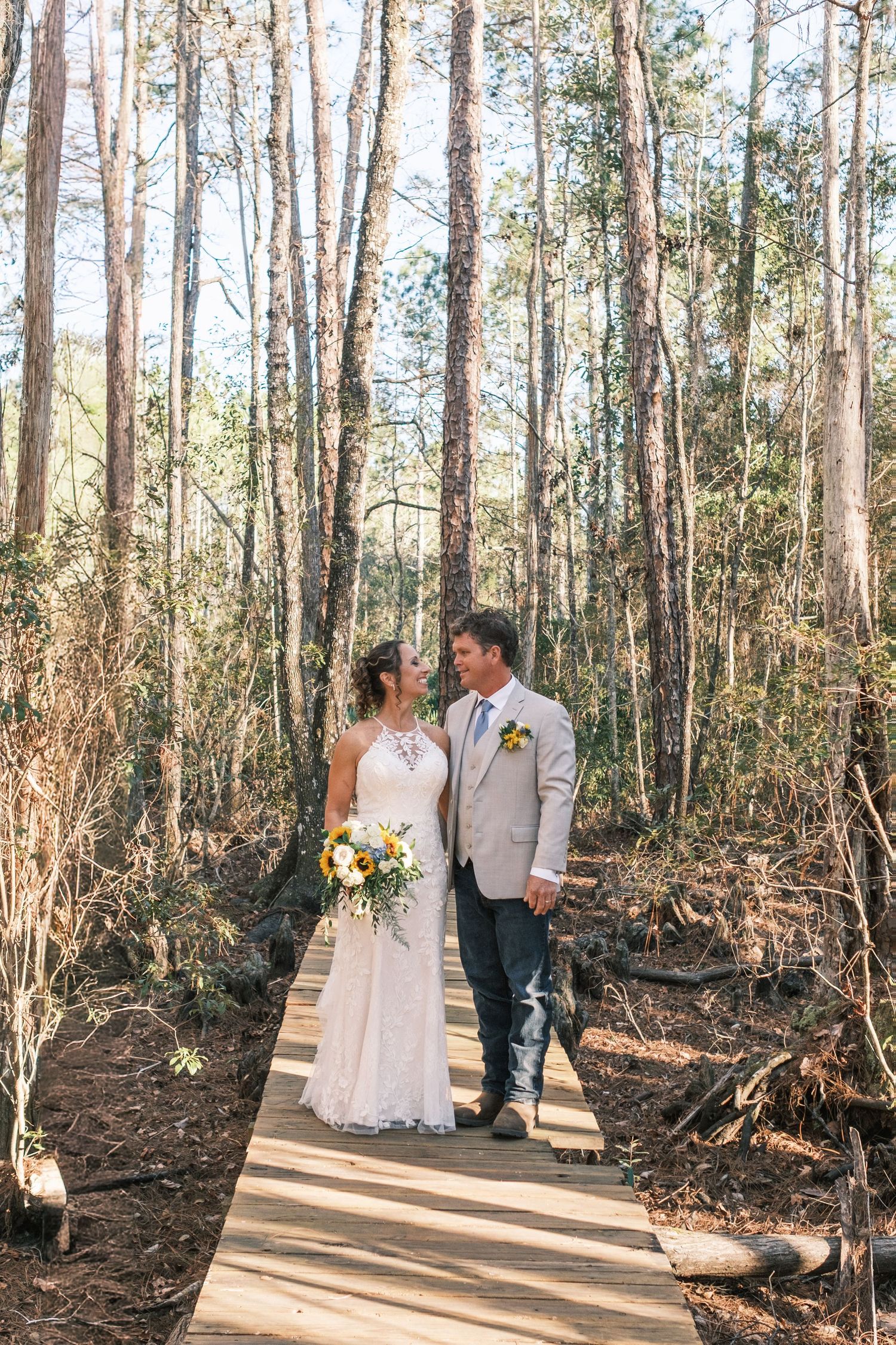 Bride and groom stand on woodland boardwalk, bride holding yellow bouquet, sharing intimate moment.