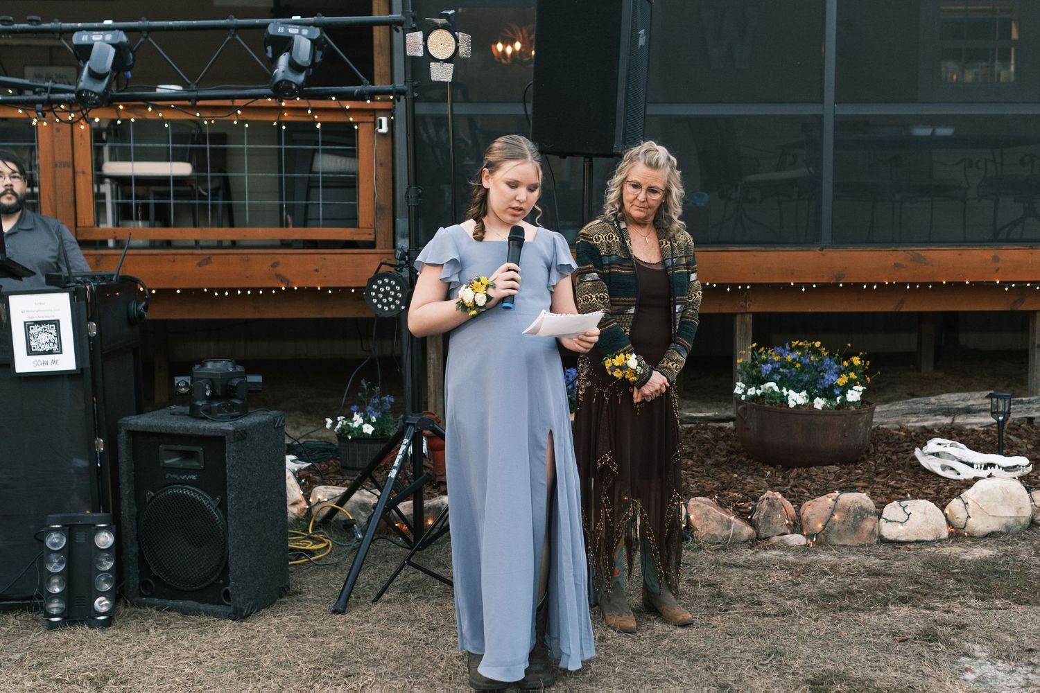 Bridesmaid in blue gown delivers a heartfelt speech at an outdoor evening wedding reception.