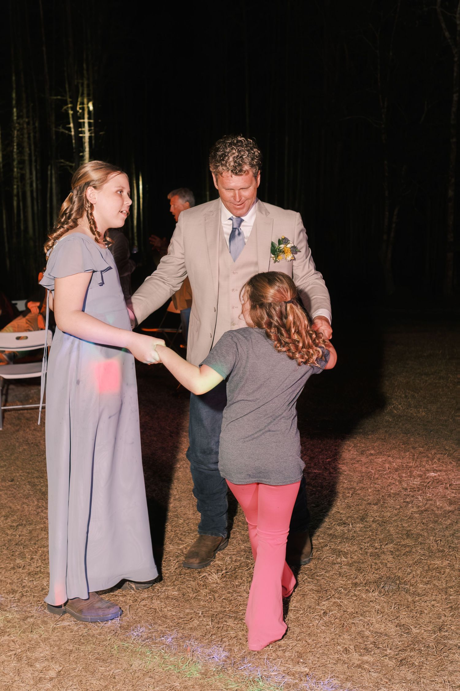 Groom in a tan suit dancing with two young girls at an outdoor evening wedding reception.