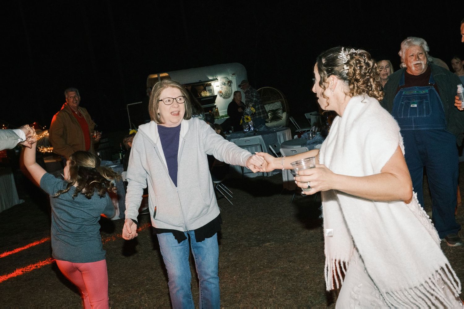 Wedding guests including bride in white shawl hold hands and dance at nighttime reception.