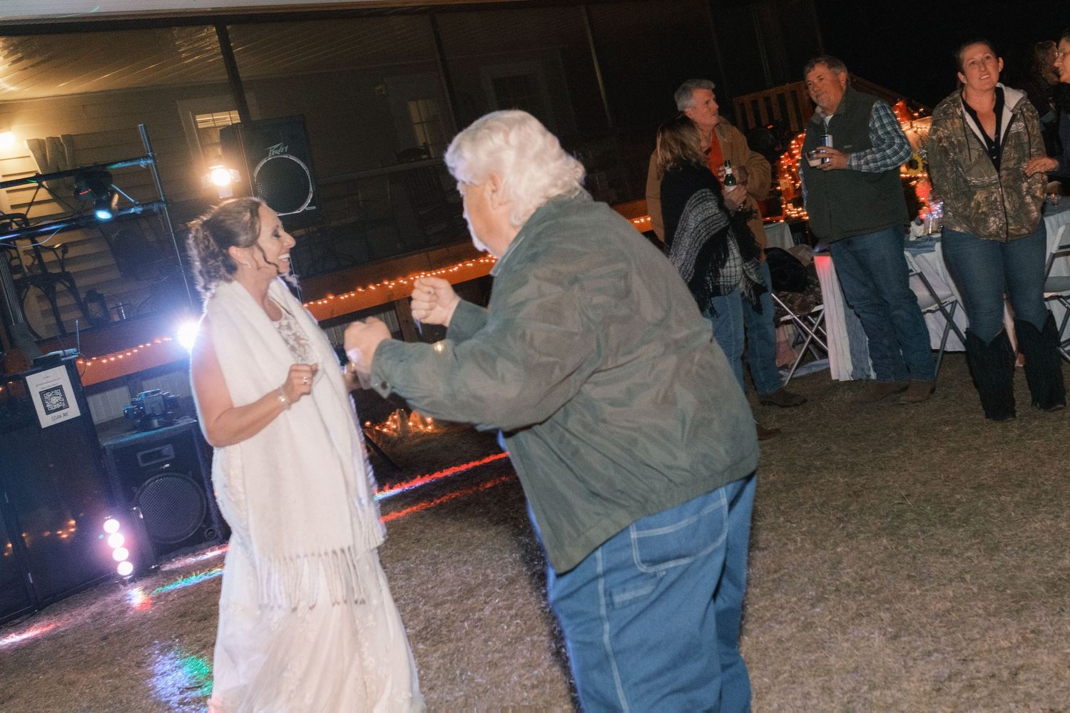 Bride in white dress dances with elderly guest at an outdoor evening wedding reception.
