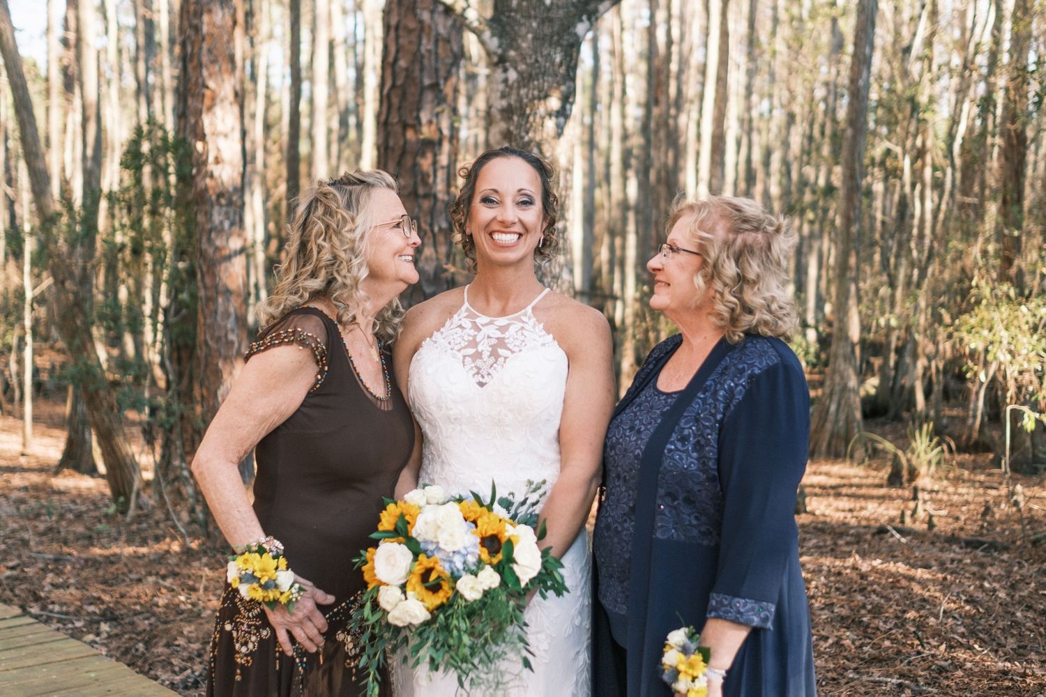 Bride in lace gown holding sunflower bouquet smiling with two women in forest setting.