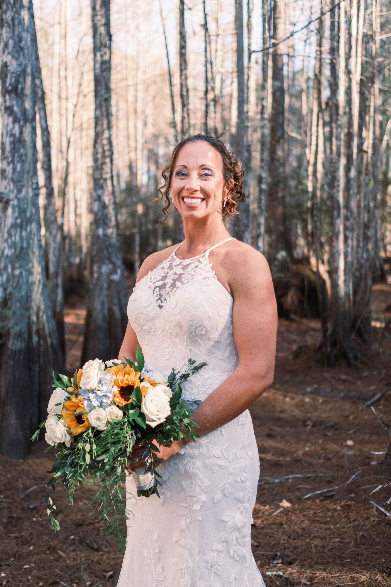 Smiling bride in lace halter dress holding yellow sunflower bouquet in wooded forest setting.
