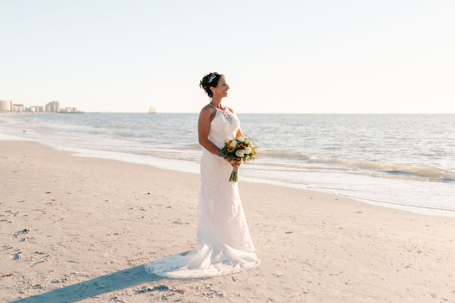 Bride in white halter gown holds colorful bouquet standing elegantly on a beach at sunset.