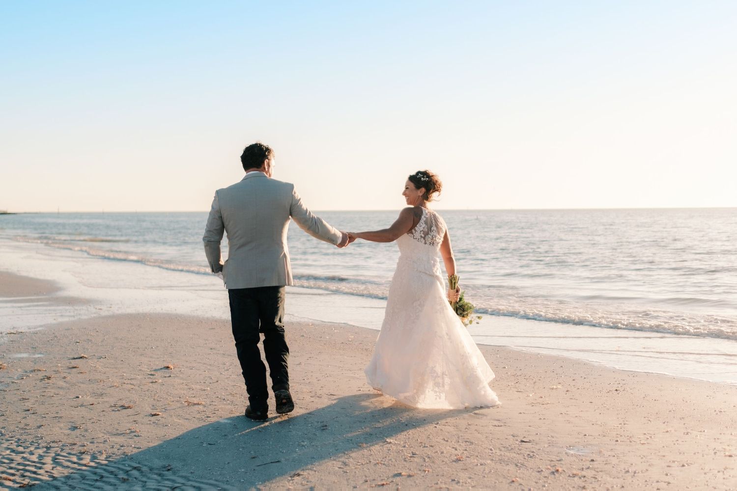 Bride and groom walk hand in hand along shoreline at sunset, bride carrying colorful bouquet.