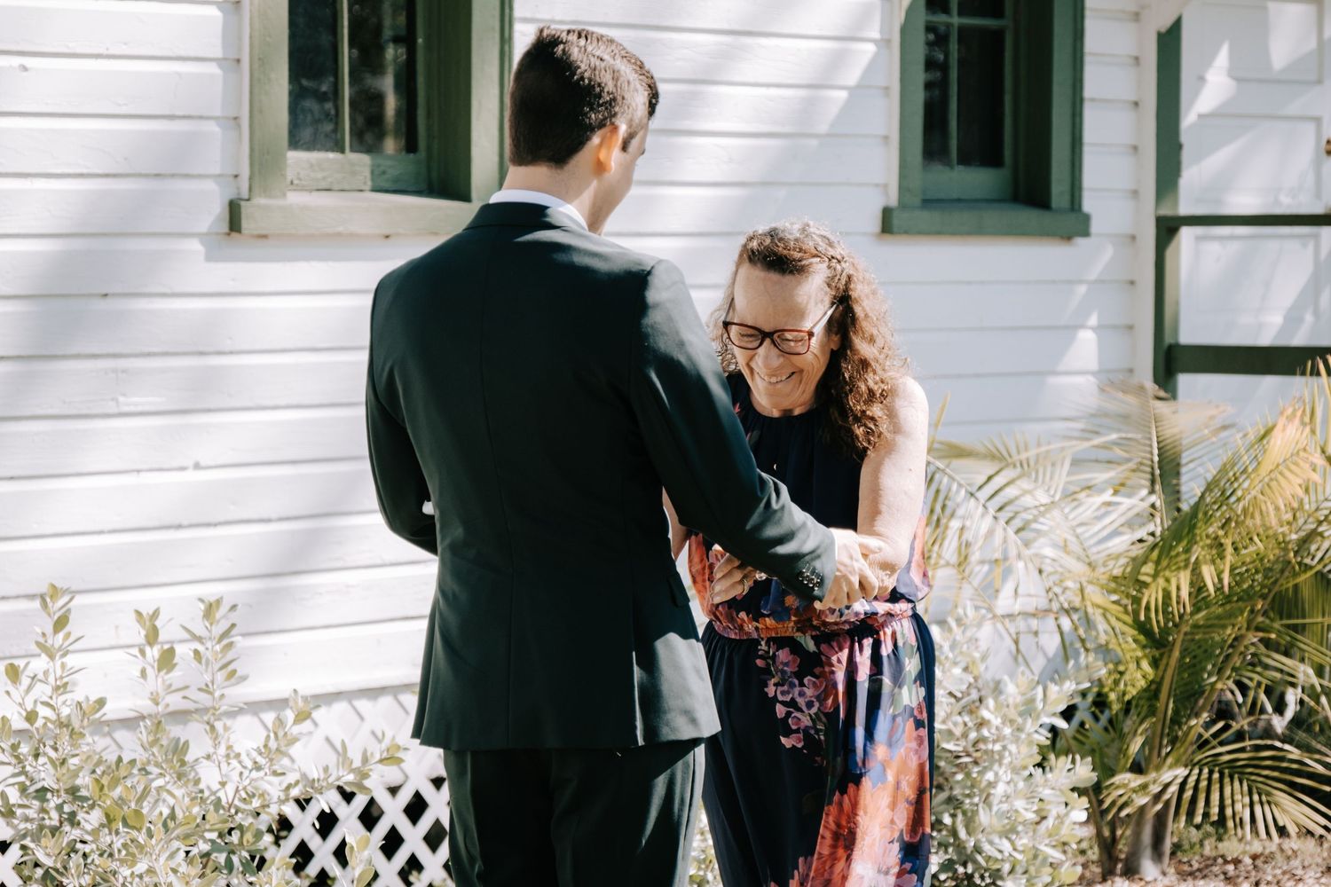A couple shares an intimate moment outside a white building with green shutters.