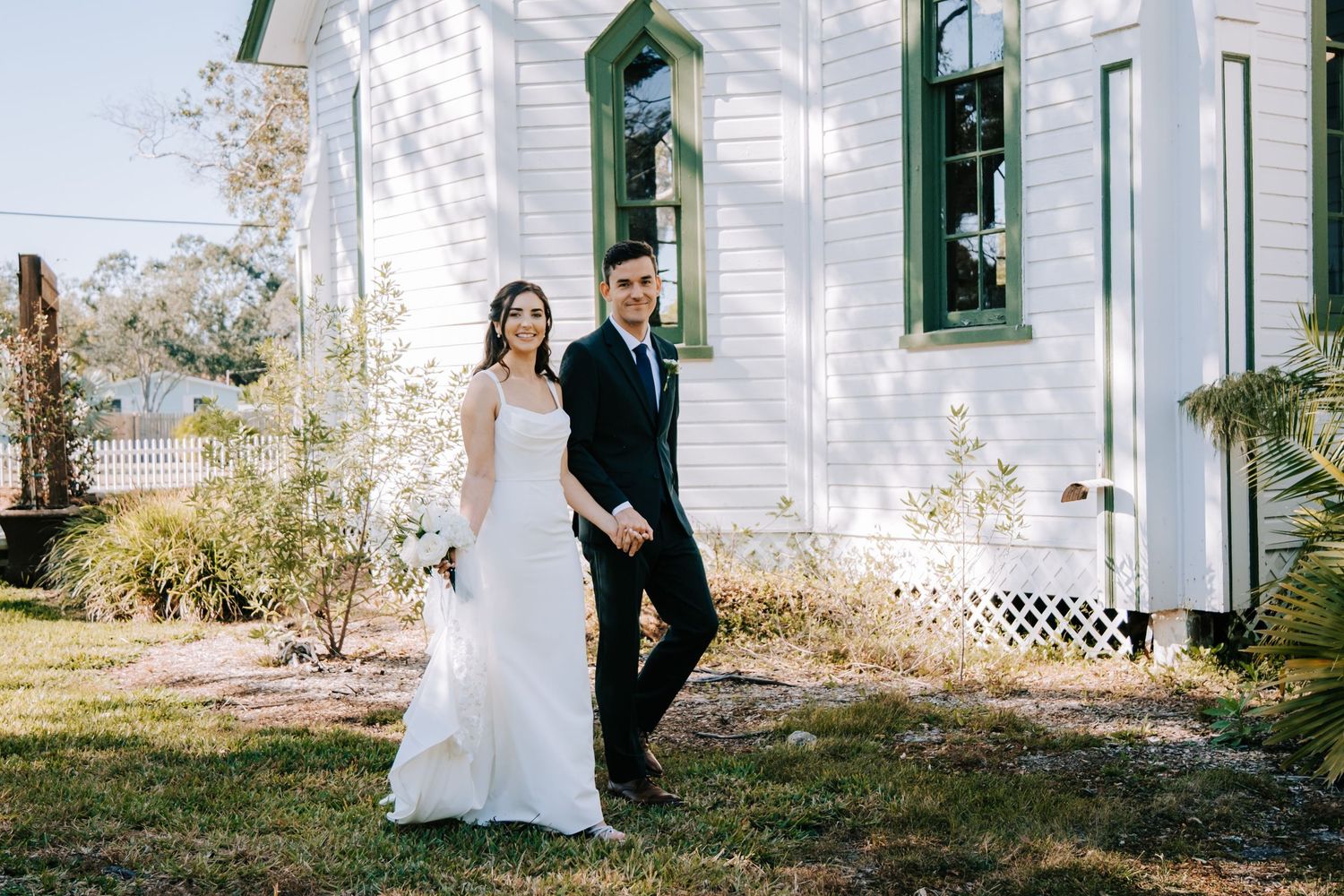 Wedding couple poses in front of a white building with green shutters and natural lighting.