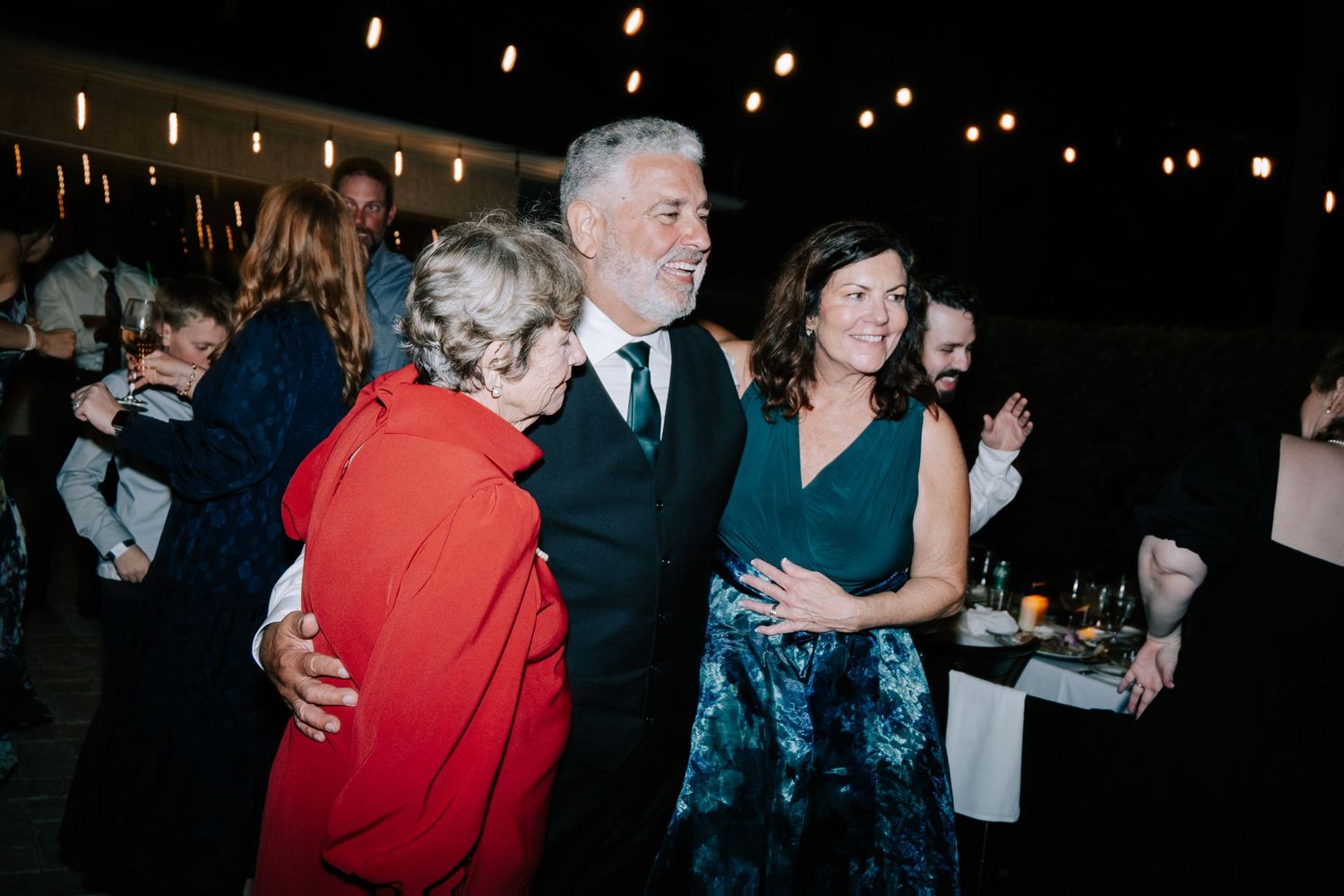 Wedding guests dancing and embracing at a dimly lit evening reception.