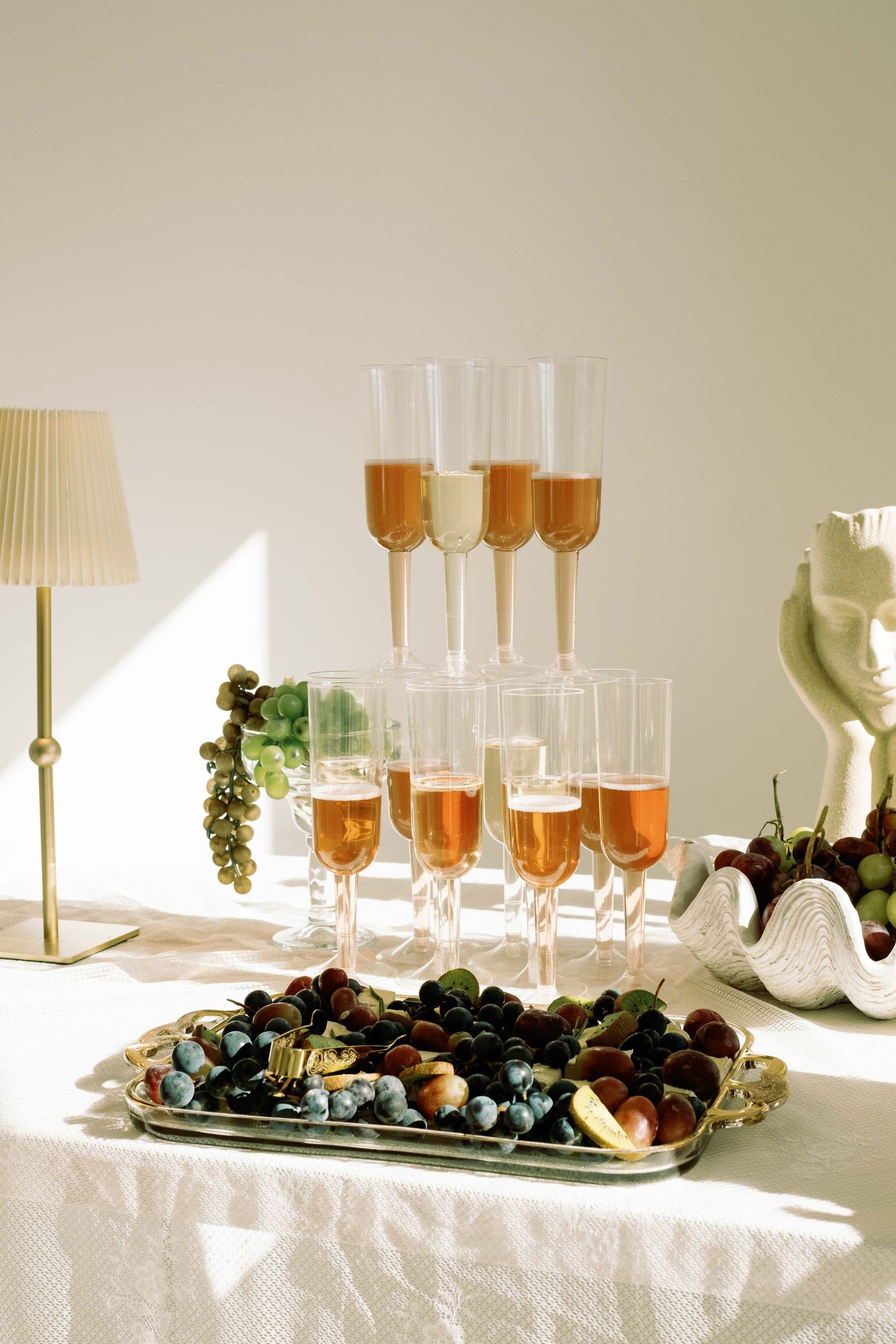 Elegant champagne tower with rose wine glasses beside a fruit platter of grapes on a white linen table.