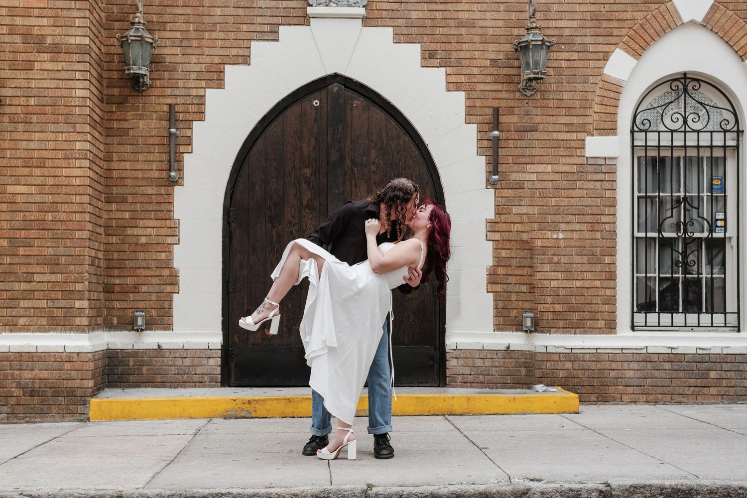 Groom dips bride in white dress outside historic brick church with arched wooden doors during wedding photo session.