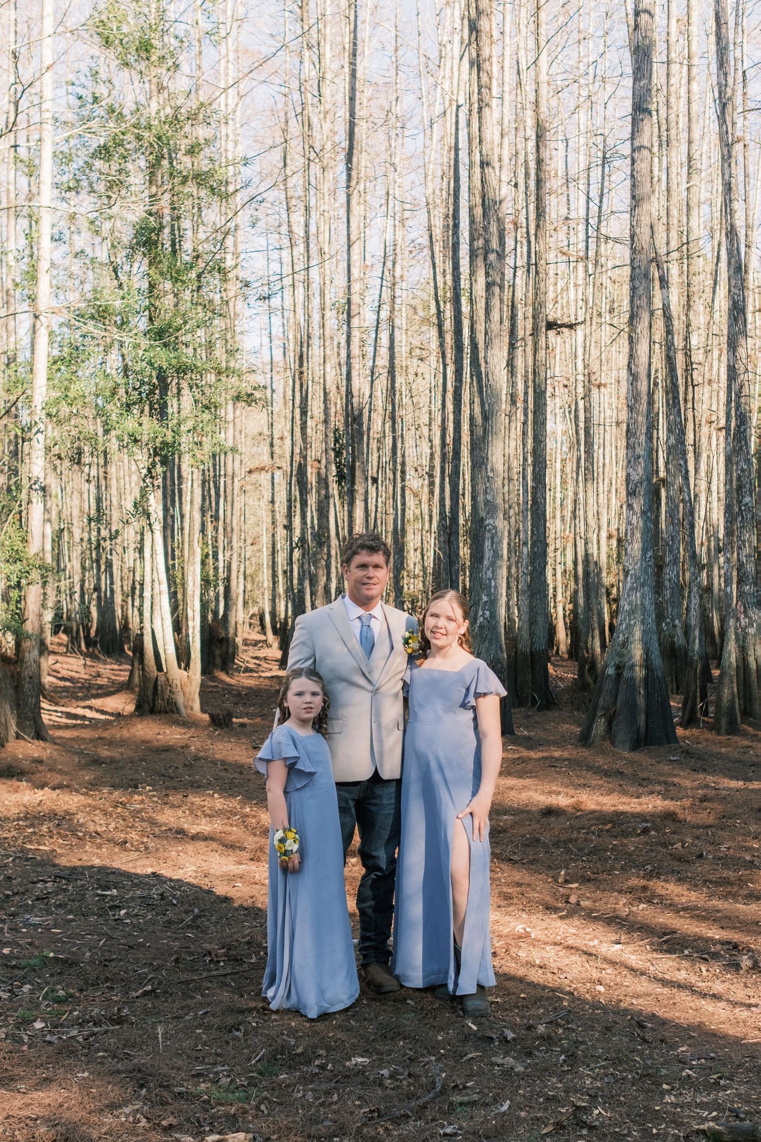 A man in a suit poses with two women in matching blue gowns among tall trees in a forest wedding setting.