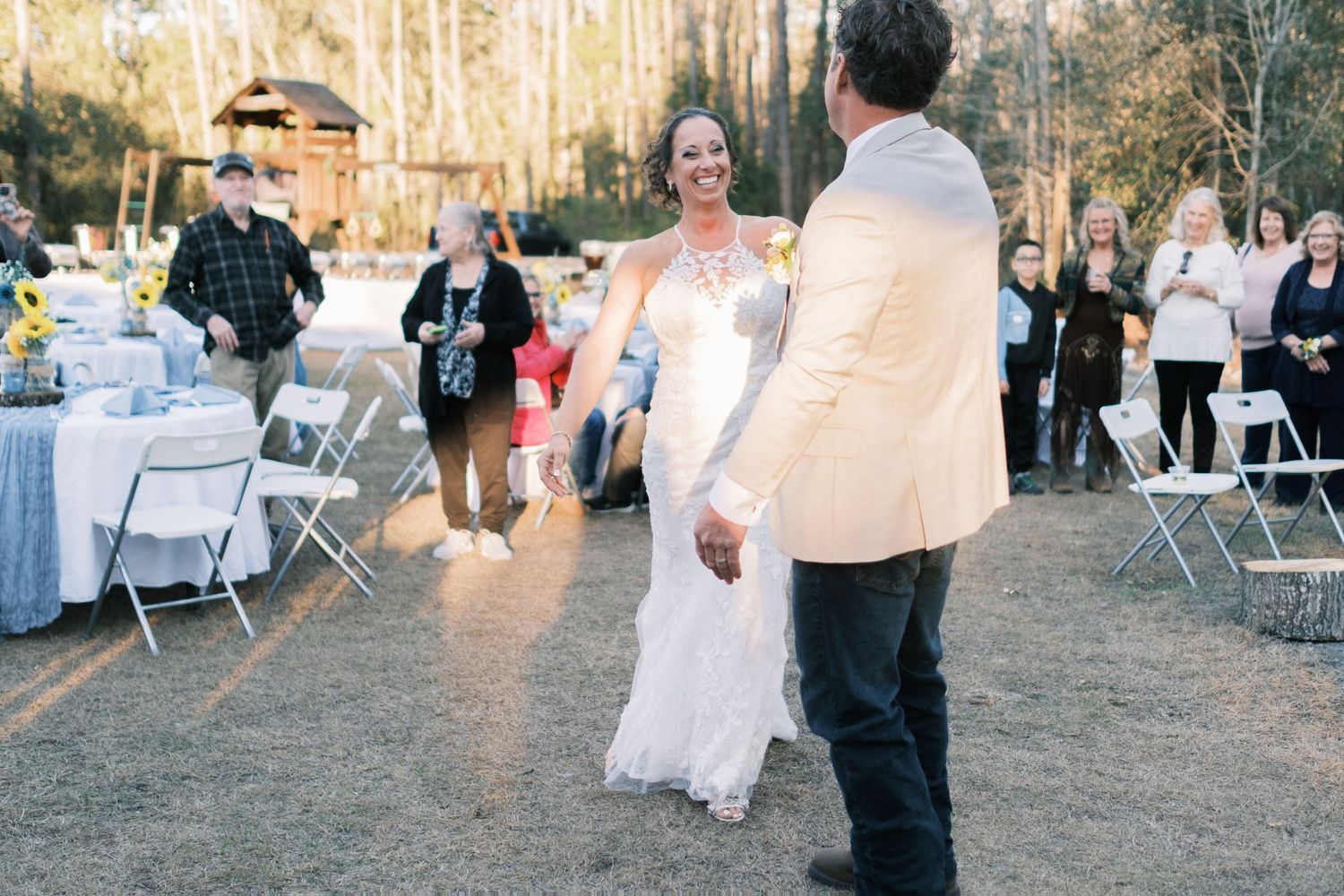 Smiling bride in lace gown dances with groom at an outdoor woodland wedding reception.