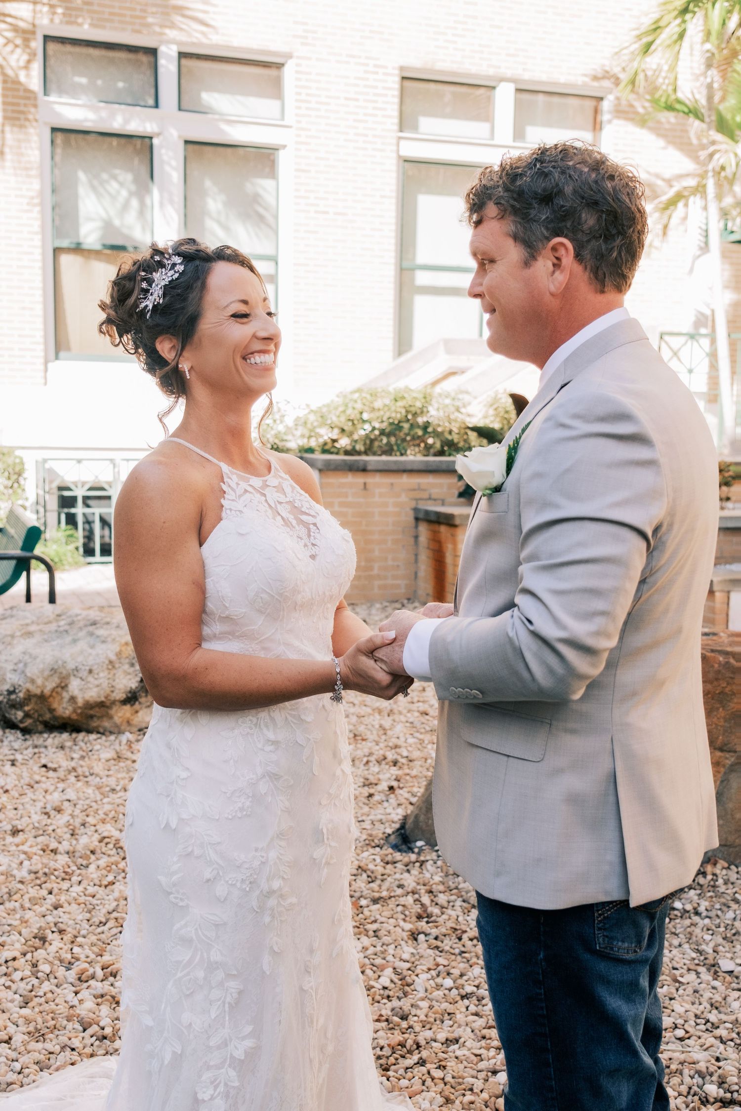 Smiling bride in lace gown holds hands with groom in tan blazer during outdoor wedding ceremony.