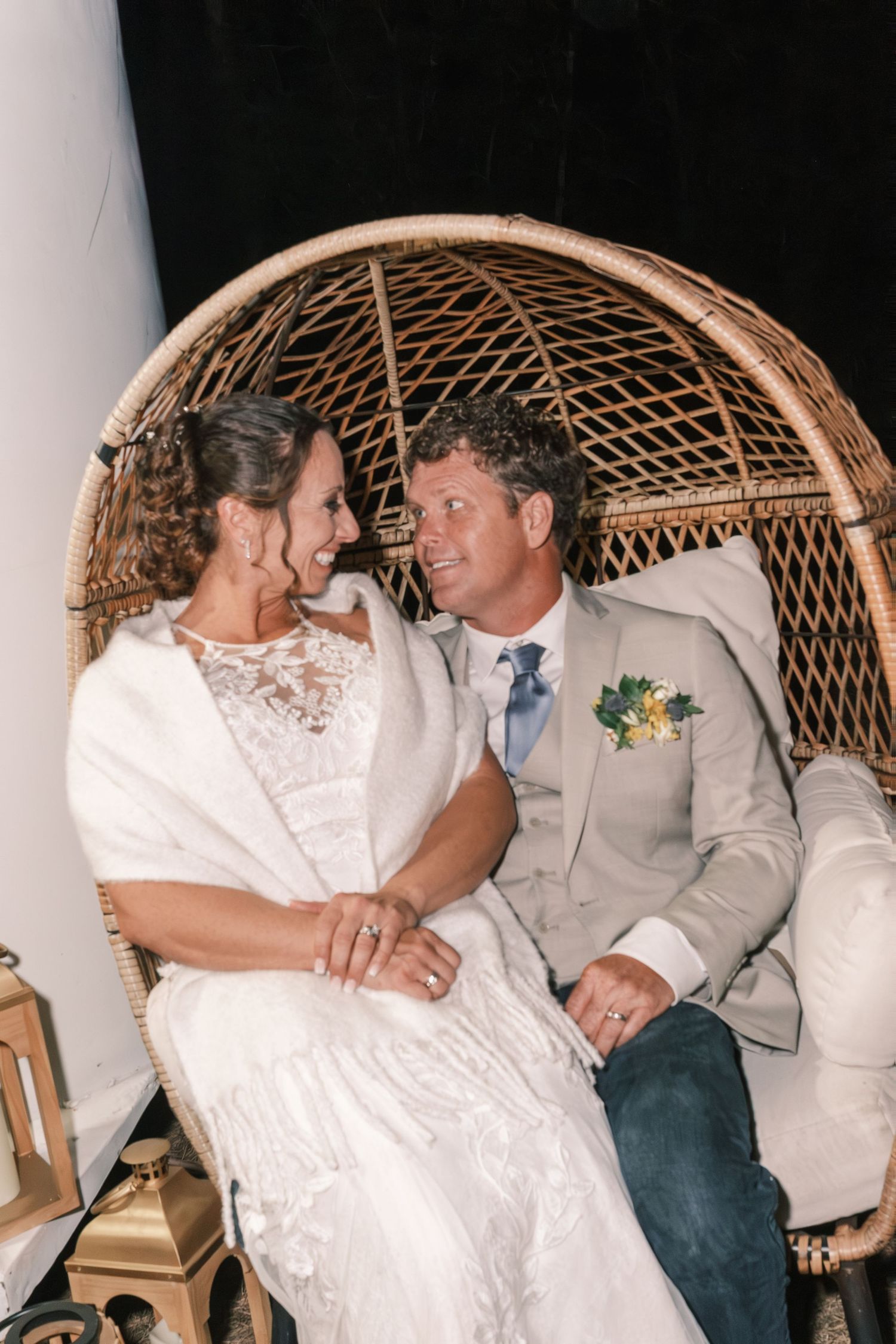 Bride and groom sit together in a large wicker egg chair, smiling at each other at night.