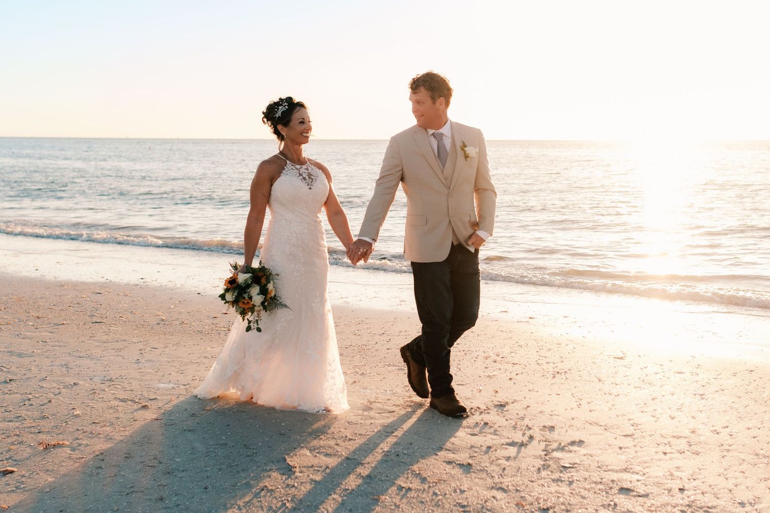 Bride and groom hold hands walking along the beach at golden hour sunset.