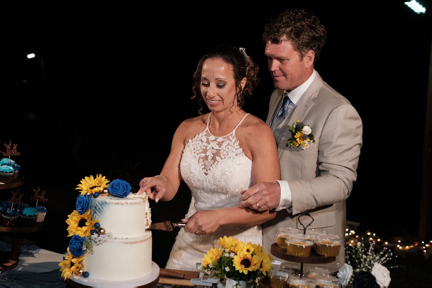 Bride and groom cutting white wedding cake decorated with sunflowers and blue roses at reception.
