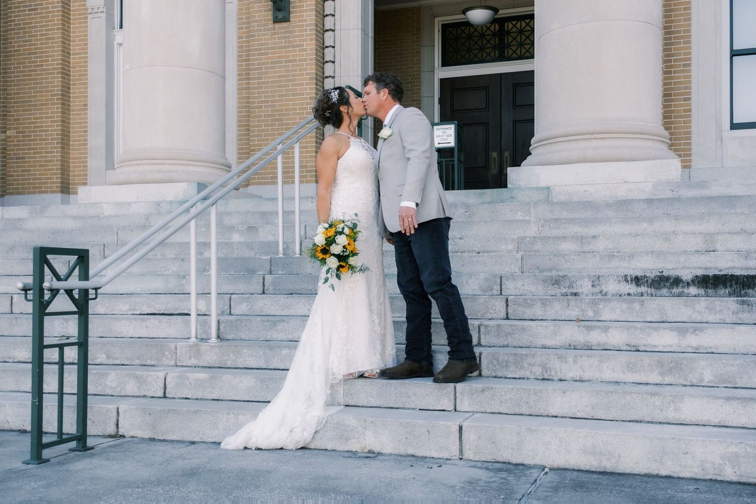 Bride and groom share a kiss on the steps of a classic brick building on their wedding day.