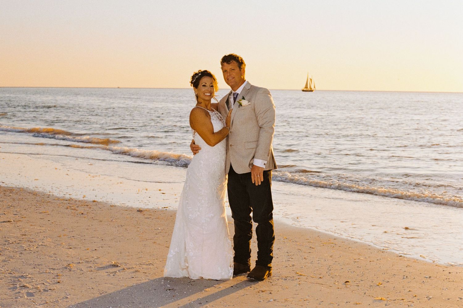 Bride and groom pose together on a sandy beach at sunset with a sailboat on the horizon.