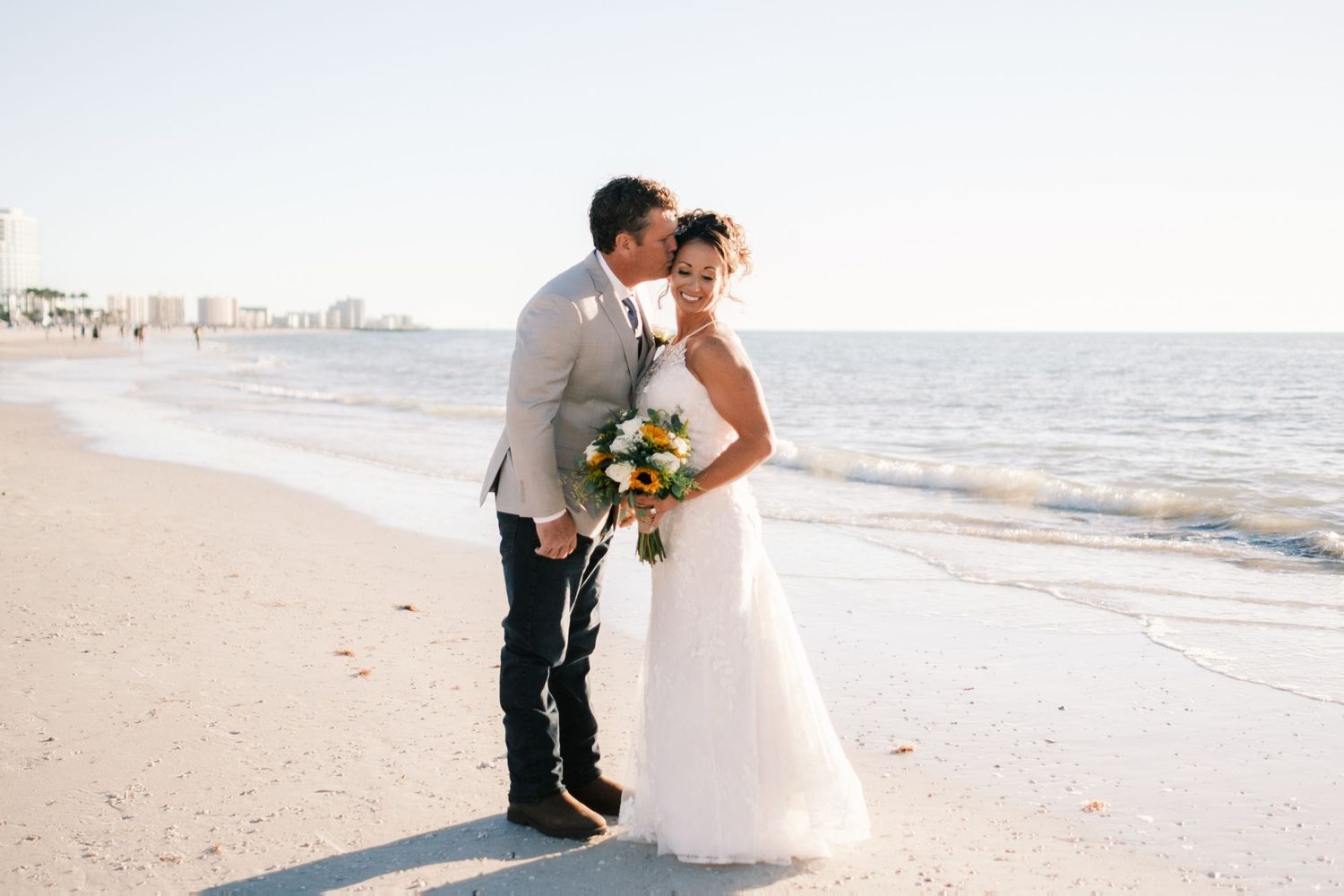 Groom kisses bride on the cheek as they stand together on a sunny beach after their wedding.