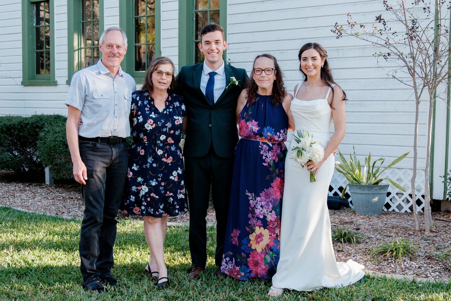 Wedding party poses for group photos outside white clapboard building.