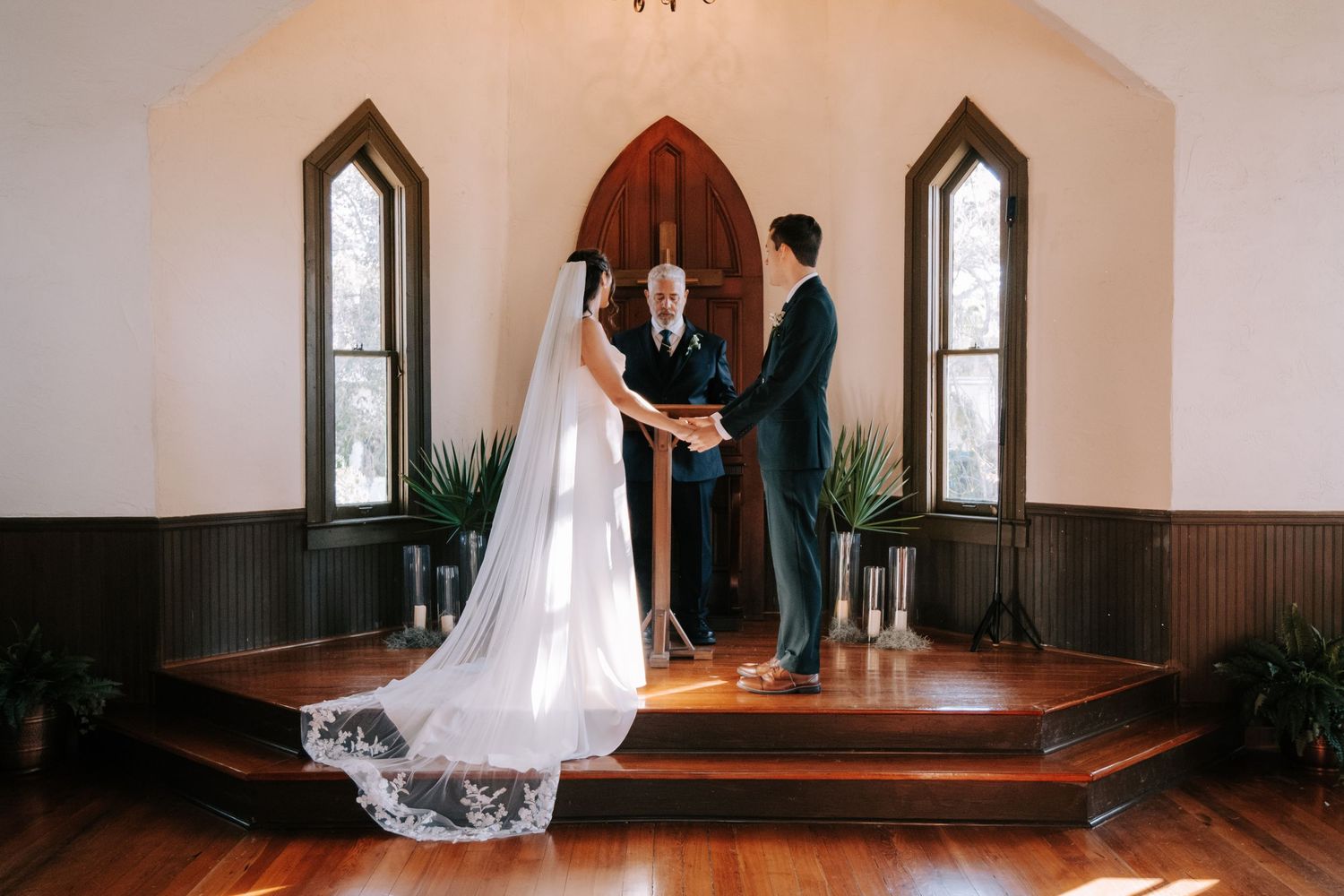 A wedding ceremony takes place in a historic chapel with Gothic arched windows and a flowing white lace veil on display.