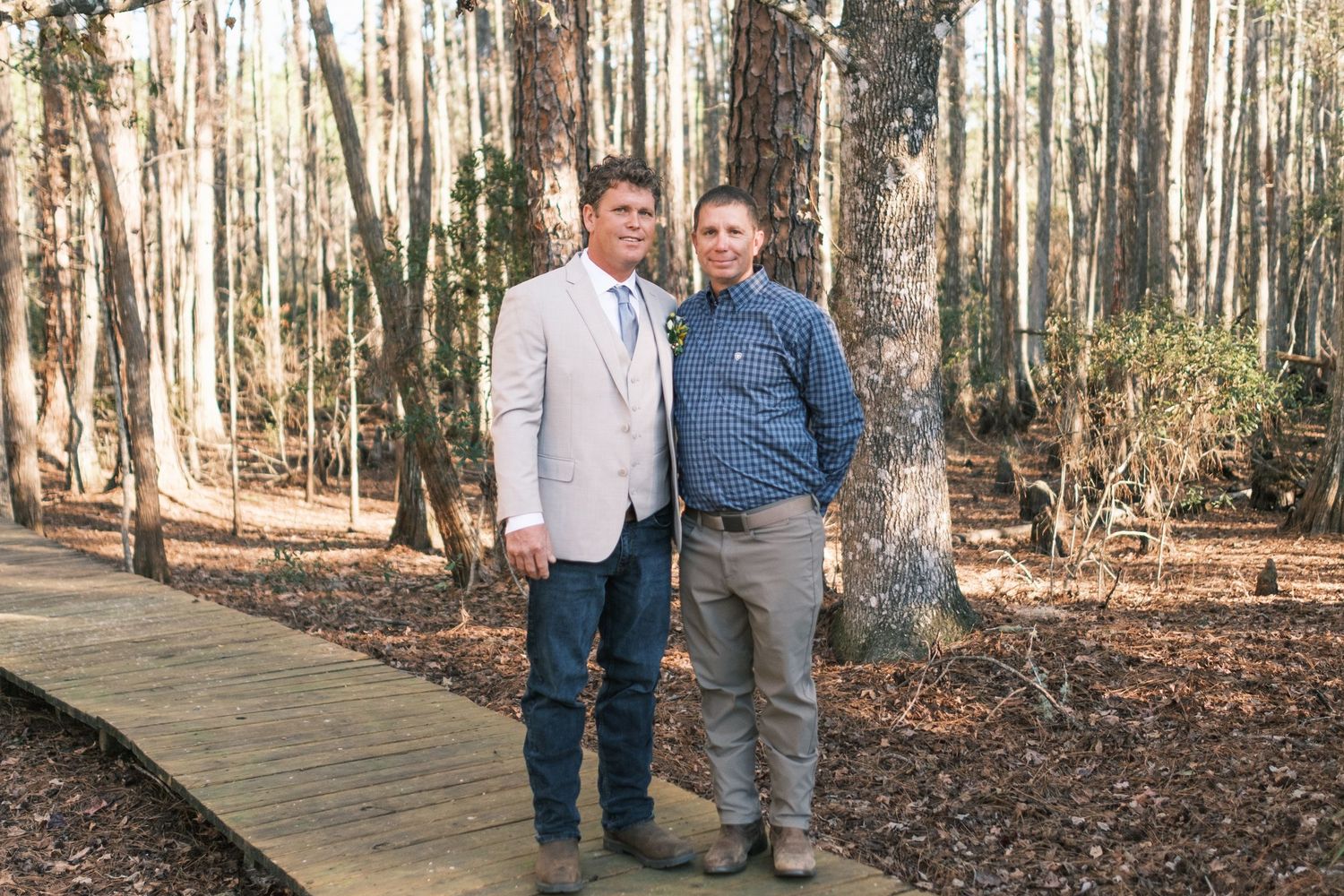 Two men in suits pose together on a wooden boardwalk surrounded by tall trees at a wedding.