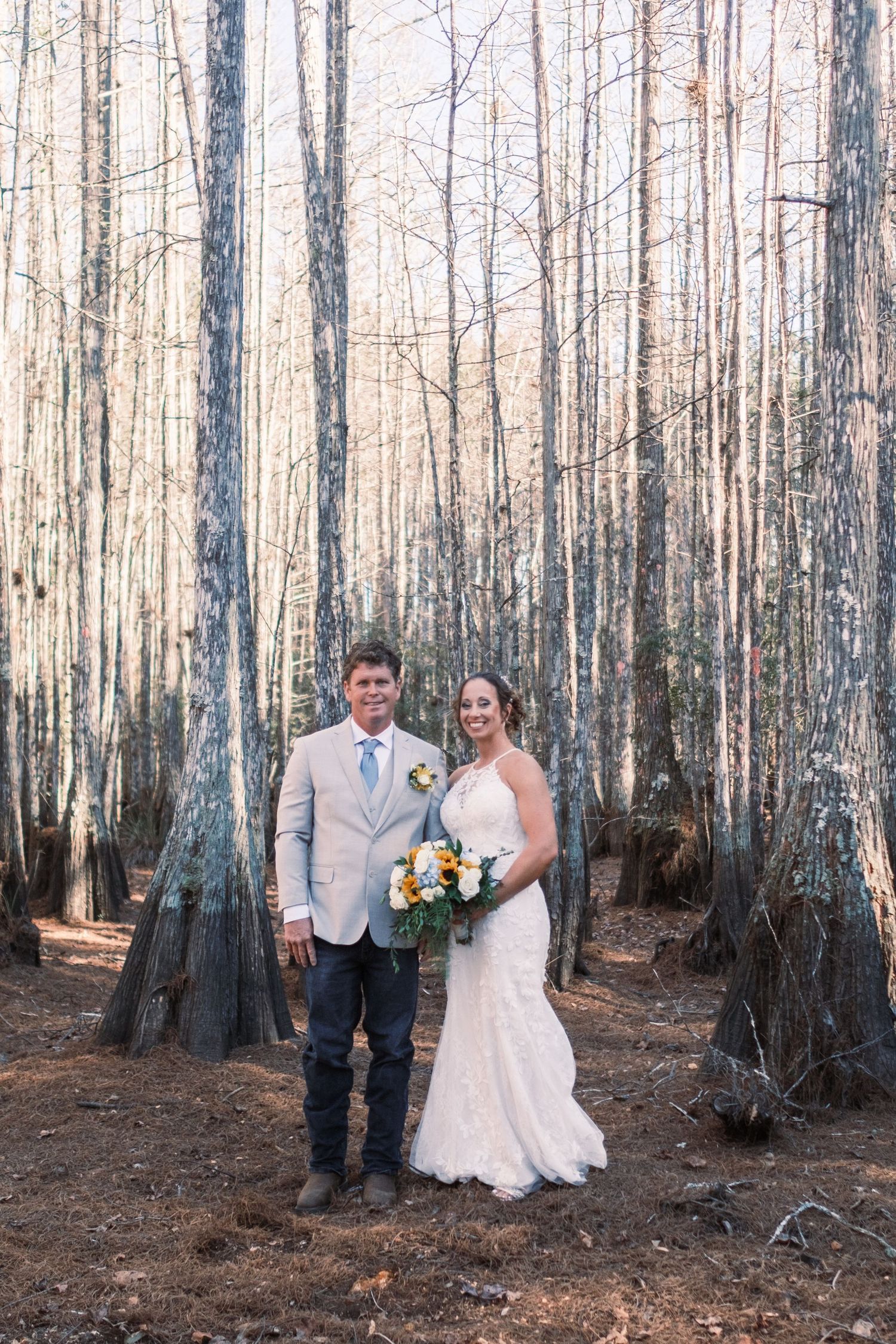 Bride and groom pose together in a rustic forest setting, bride holding sunflower bouquet in lace gown.