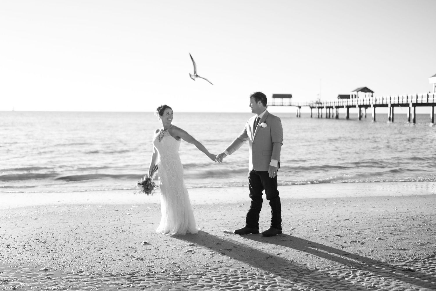 Black and white photo of bride and groom holding hands on a sandy beach with a pier and seagull in background.