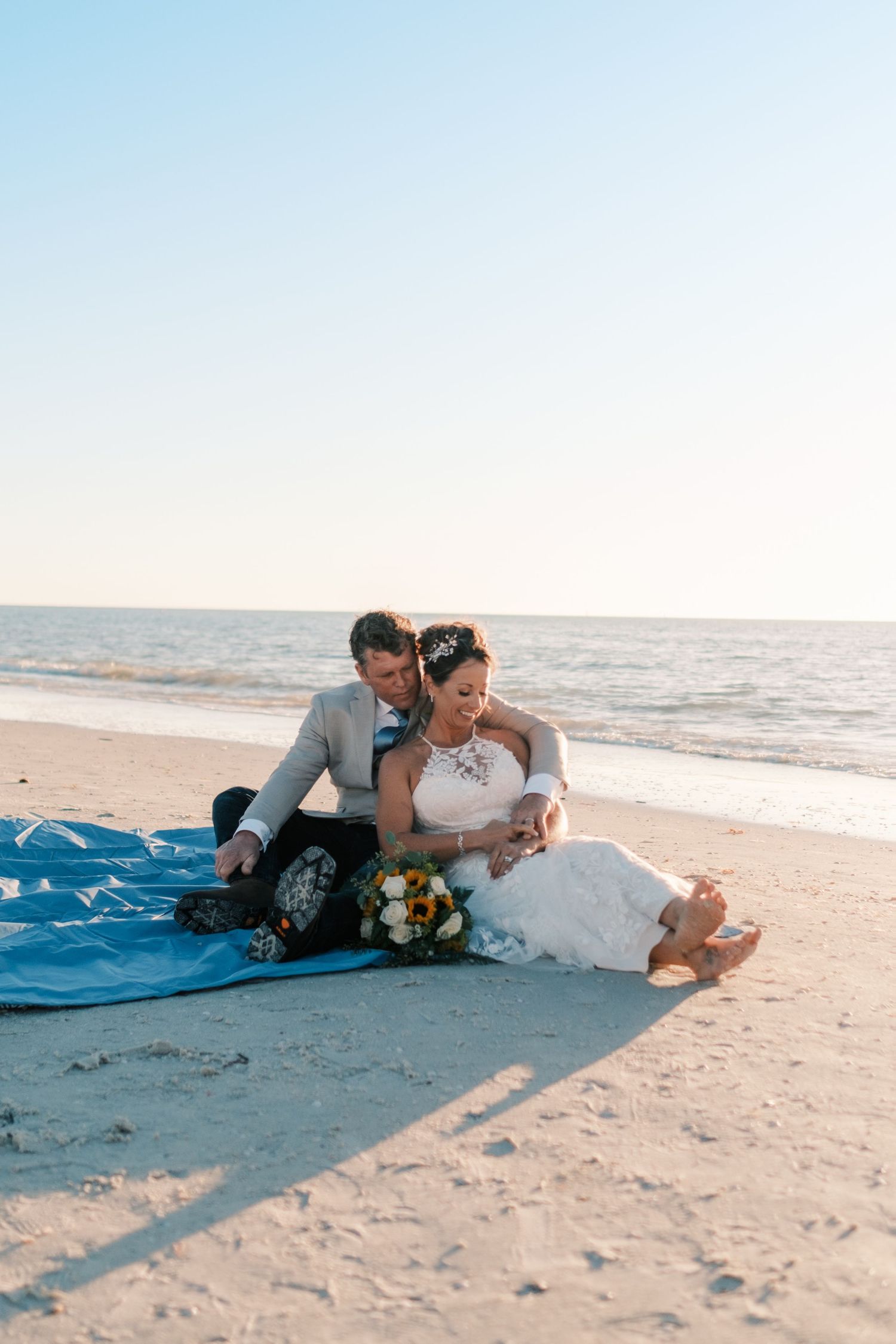 Bride and groom sit together on a blanket on the beach, sharing an intimate moment at sunset.
