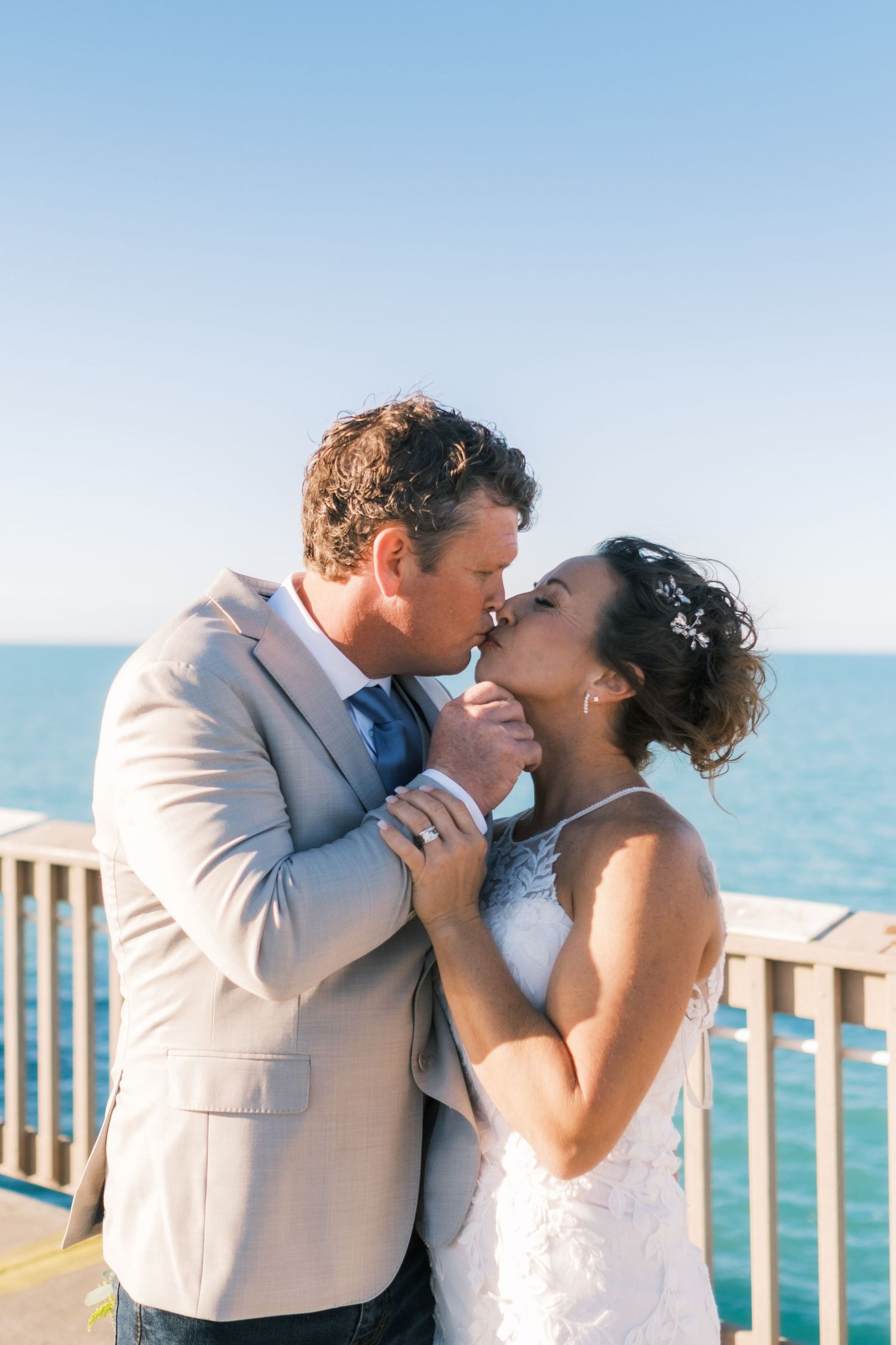 Groom in tan suit kisses bride in white lace dress on seaside pier with blue ocean backdrop.