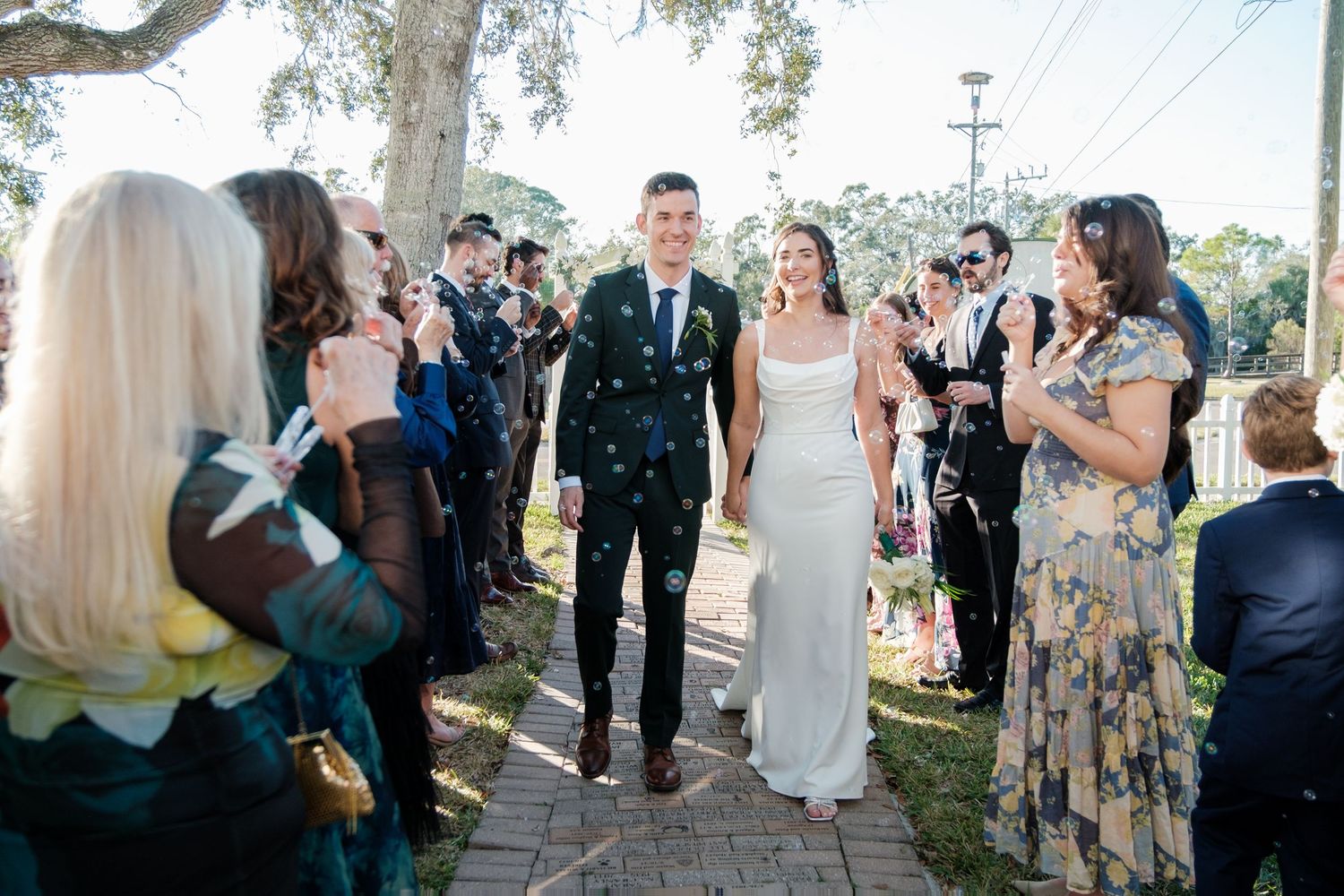 Outdoor wedding celebration with guests throwing confetti under tree canopy.
