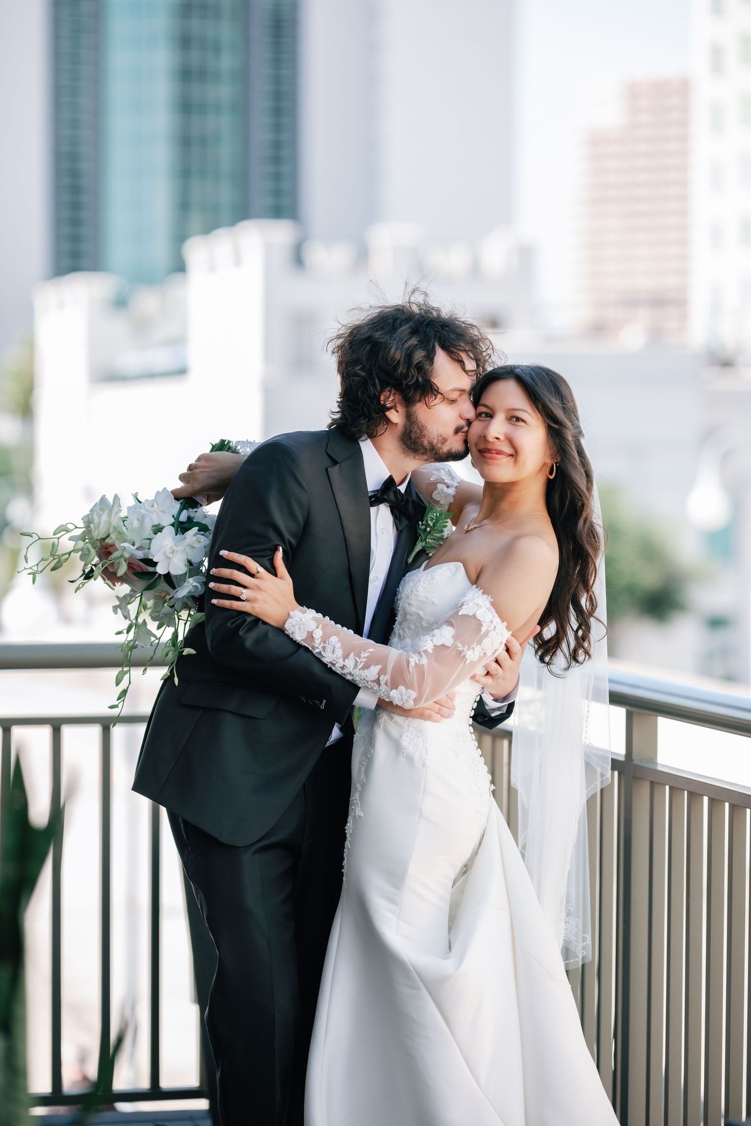 A groom in a black tuxedo kisses his bride in a white lace gown on a rooftop balcony with a city skyline backdrop.