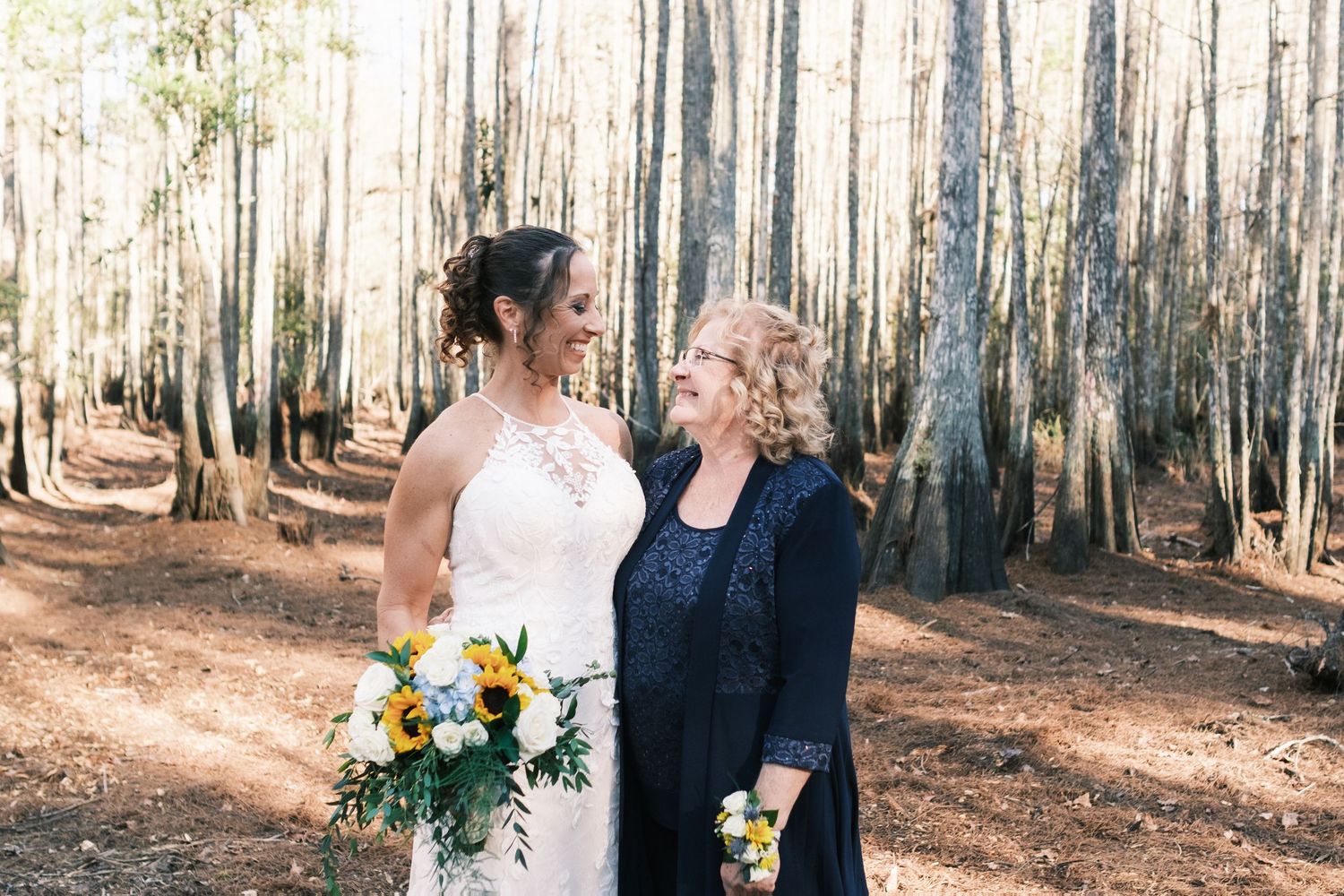Bride in lace halter gown holds sunflower bouquet beside older woman in navy dress in pine forest.