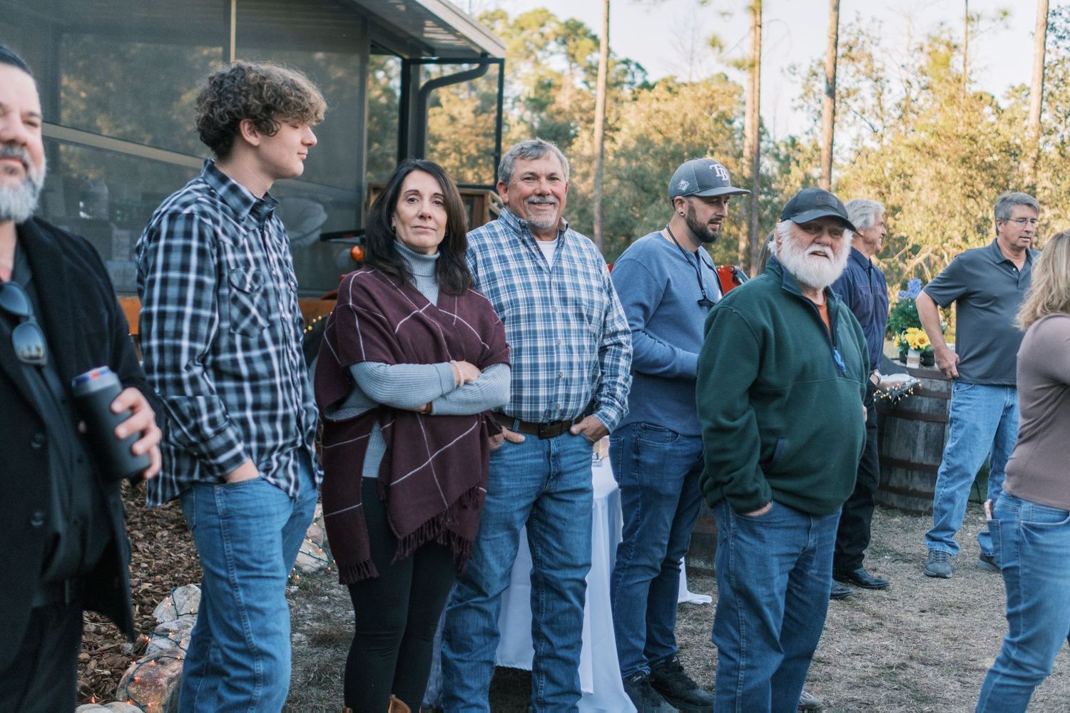 Group of guests socializing outdoors at a casual evening event near a rustic building surrounded by trees.