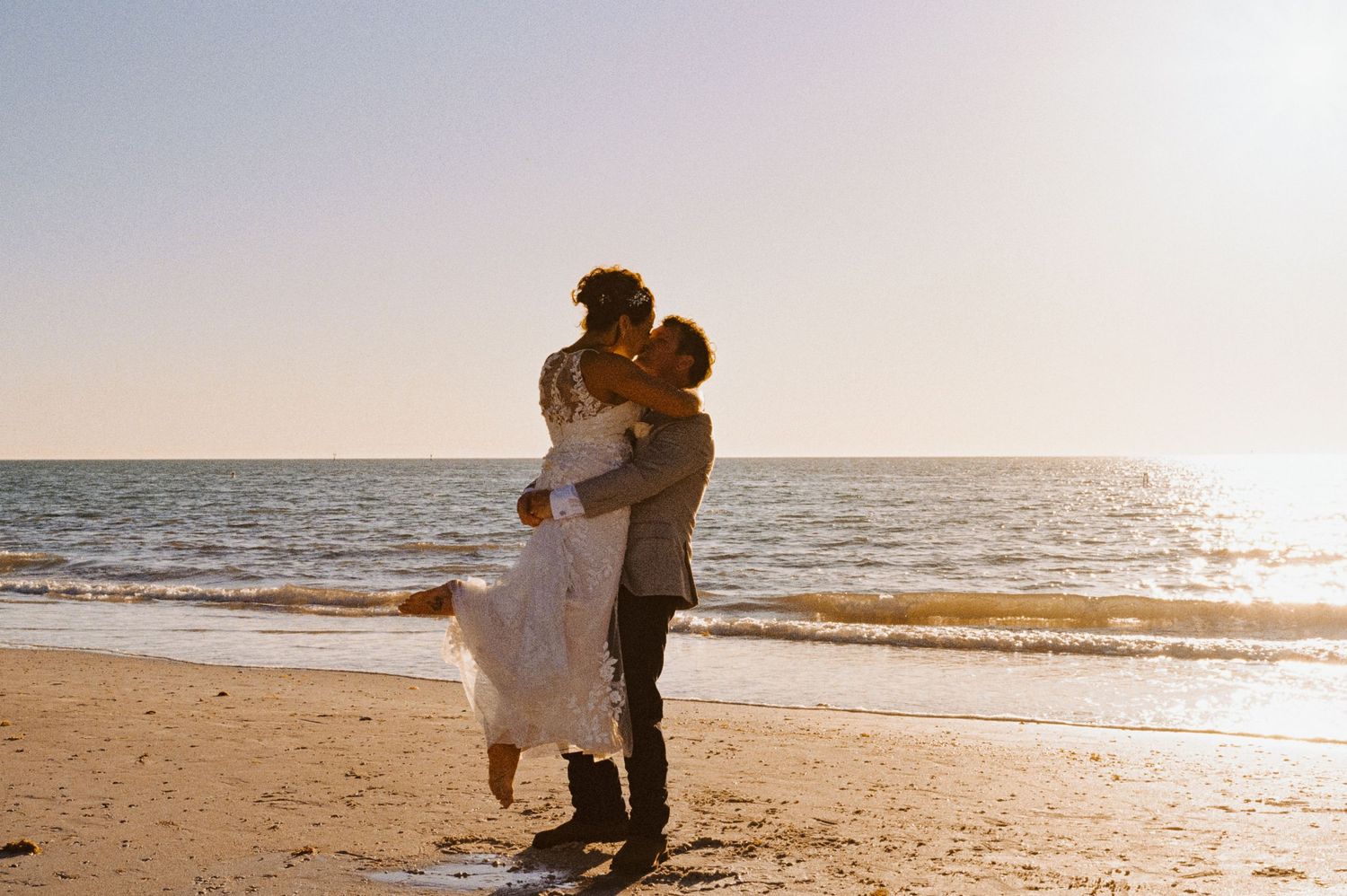 Groom lifts bride in lace gown for romantic kiss on sandy beach at golden sunset.