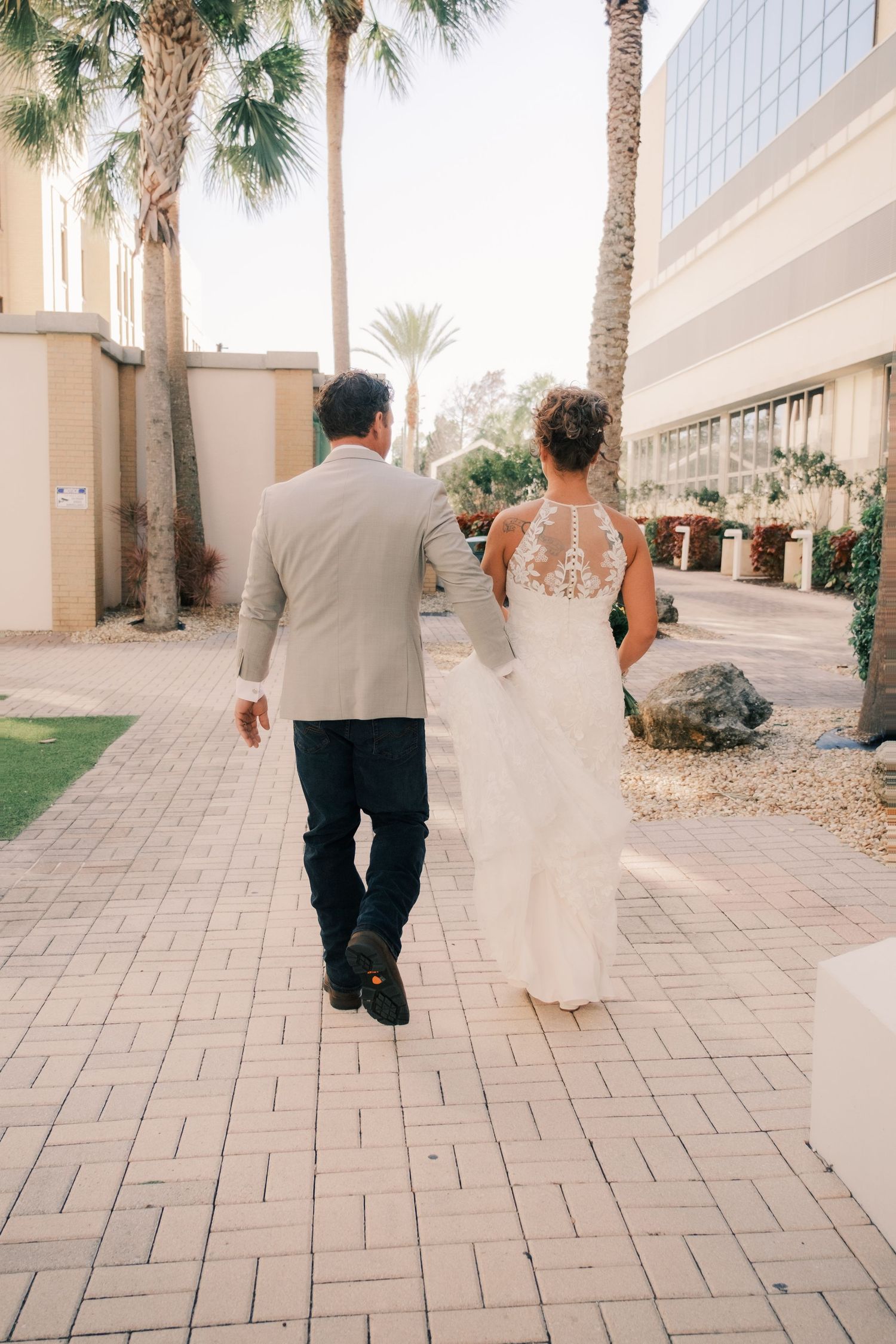 Bride and groom walking hand in hand along a palm tree-lined brick pathway outside a wedding venue.