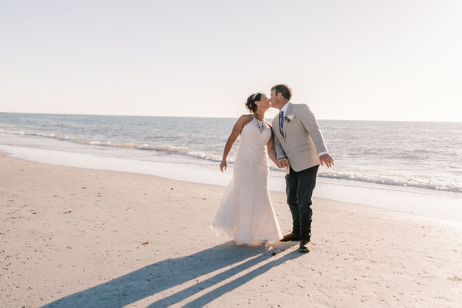Bride and groom share a romantic kiss on the beach with the ocean glowing behind them.