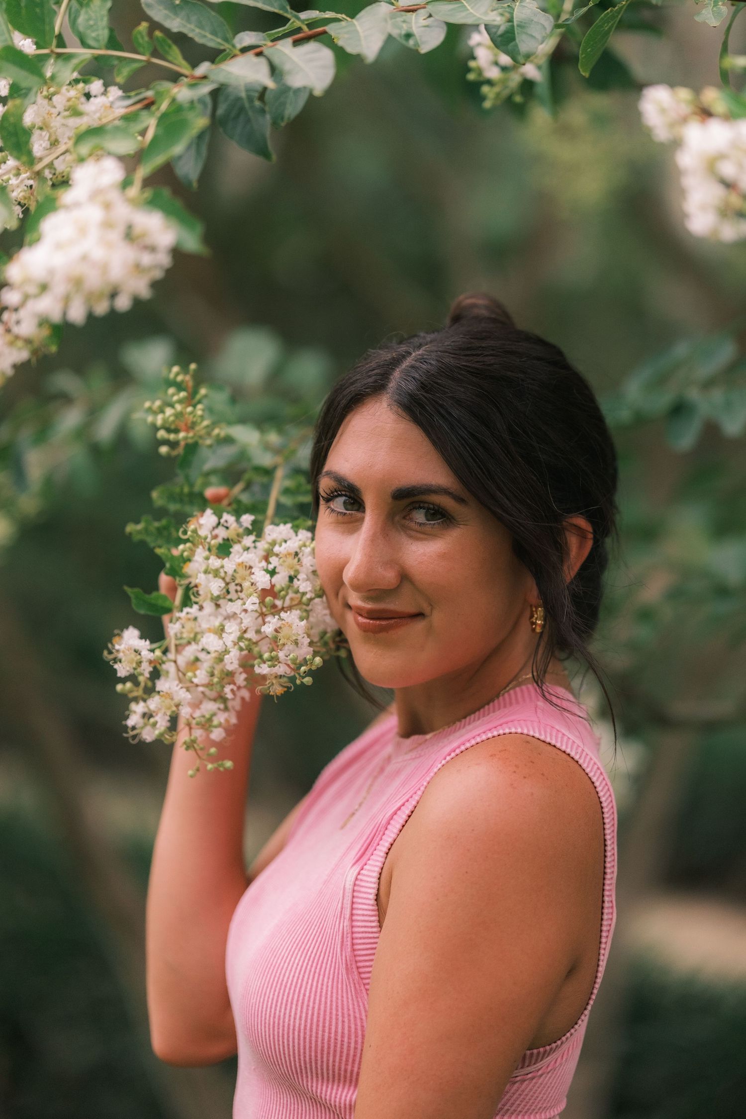 Woman in pink tank top poses with white flowering branches in a garden setting.