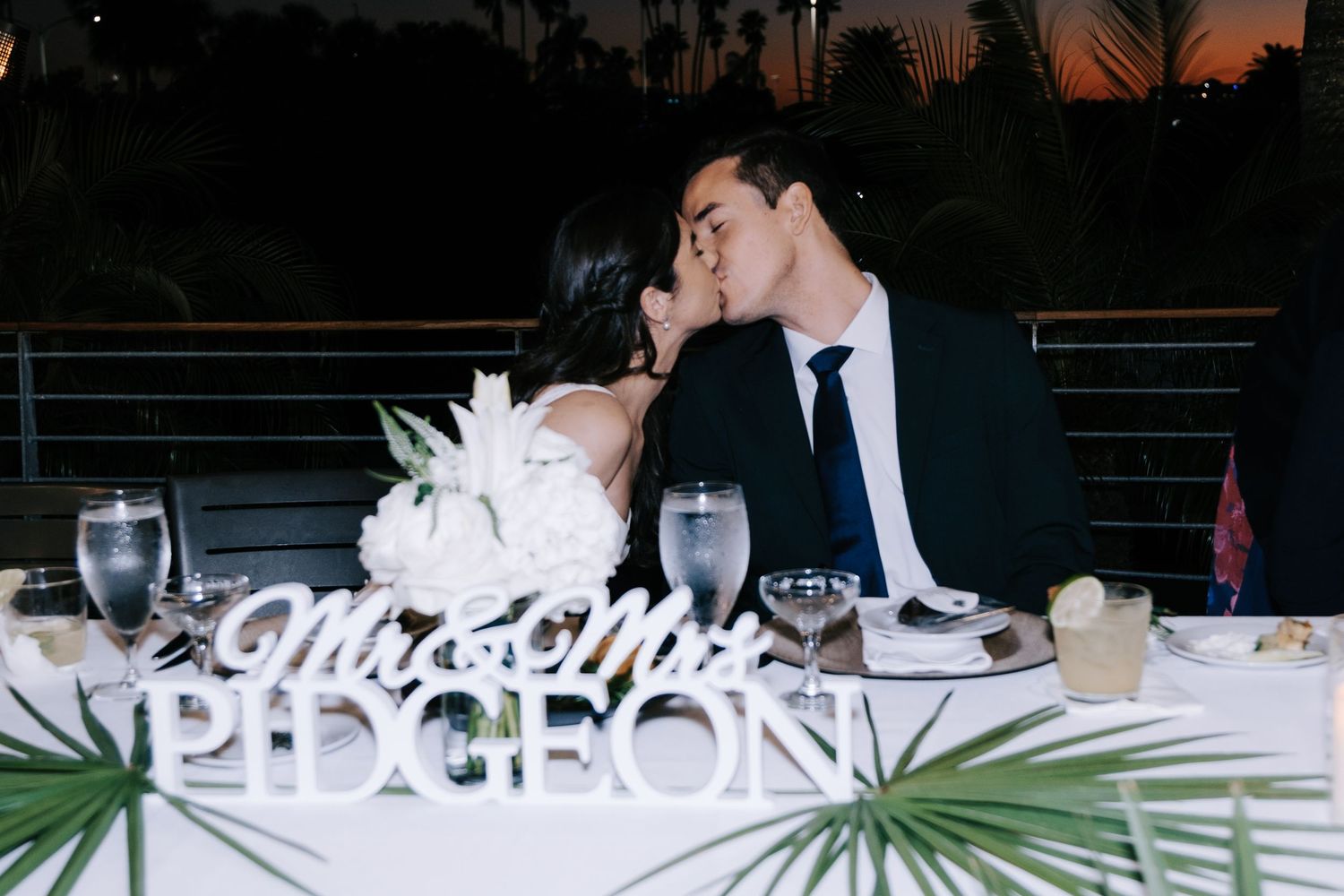 Wedding reception table setup with string lights and palm decor at sunset.