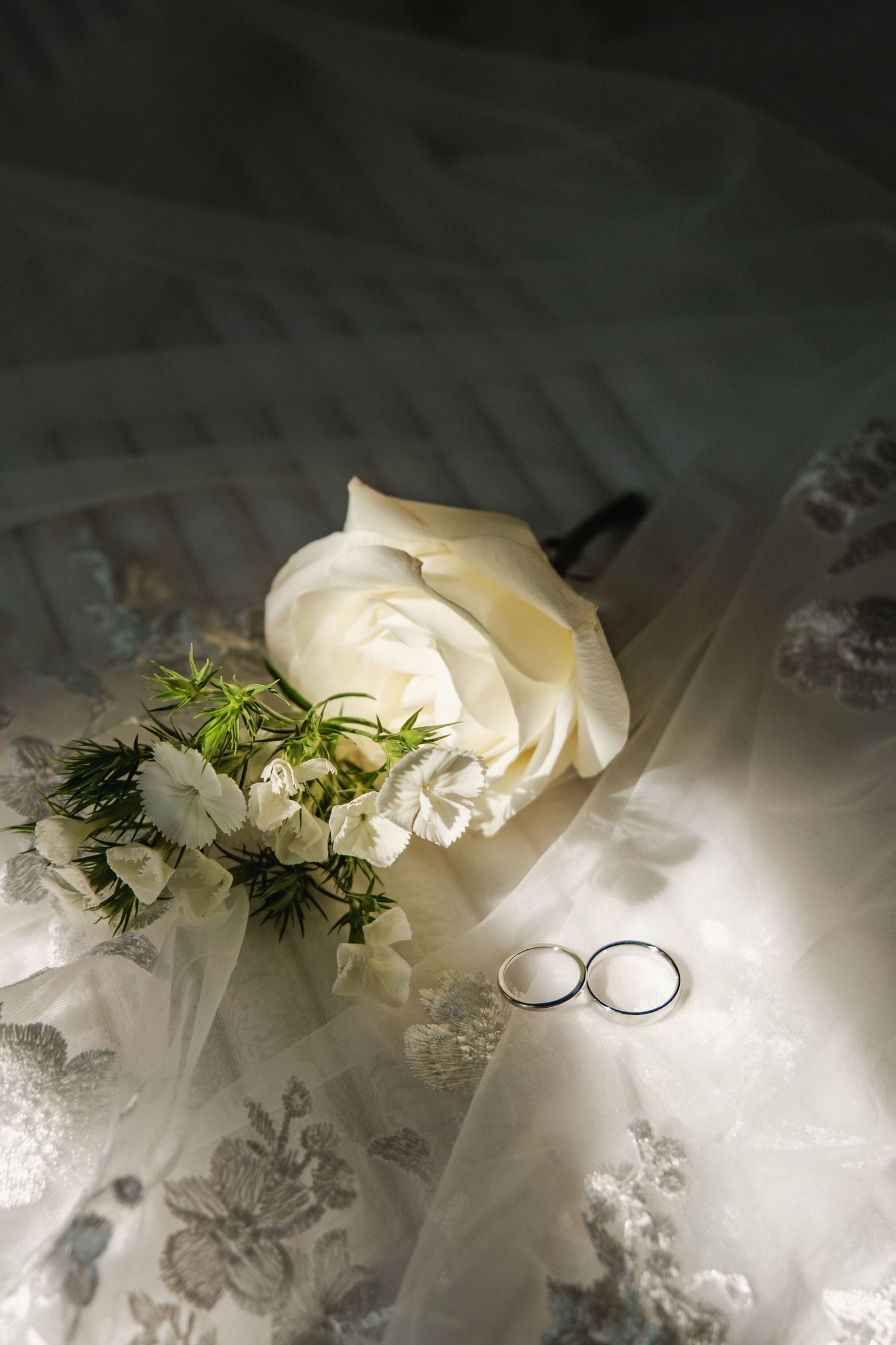 White roses and wedding rings arranged on delicate lace fabric with soft lighting.