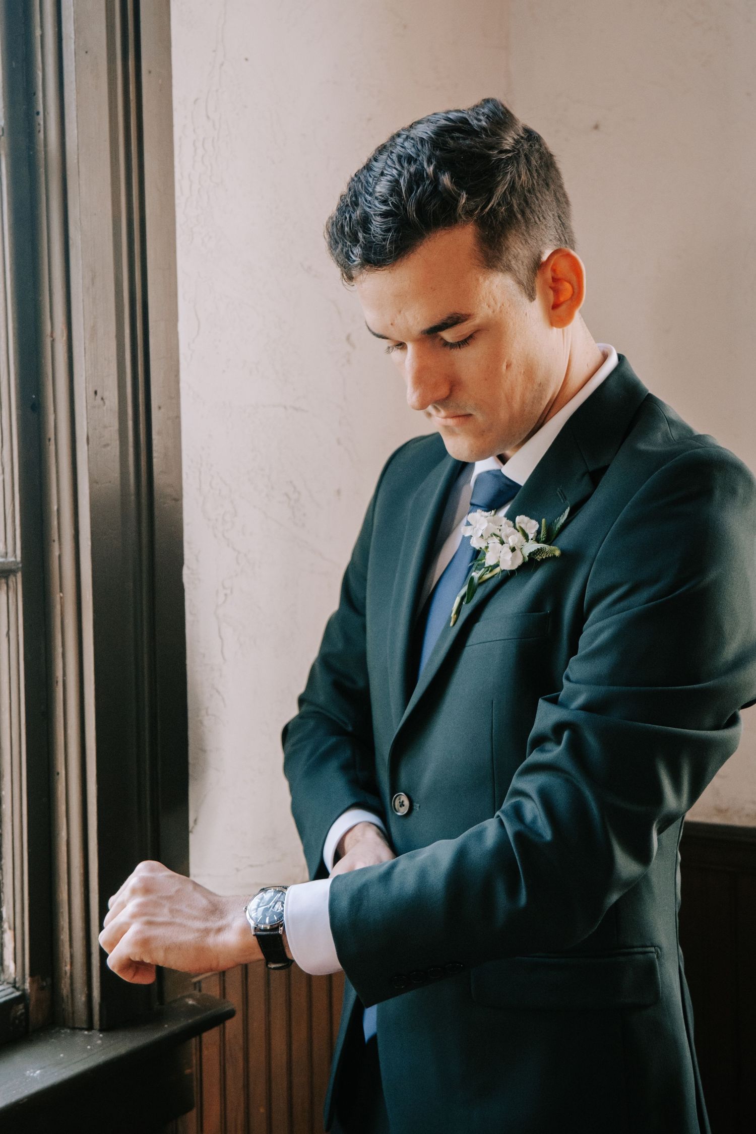 A groom in a navy suit checks his watch while standing by a window.