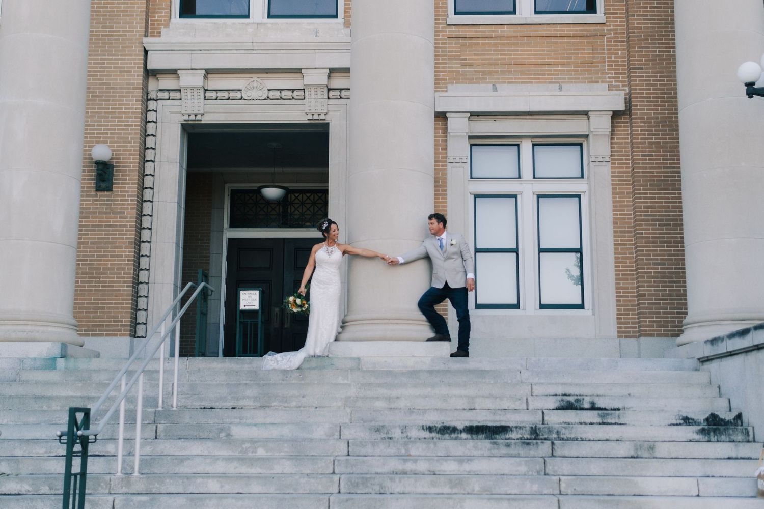Bride and groom hold hands on grand stone steps outside a classic brick building on their wedding day.