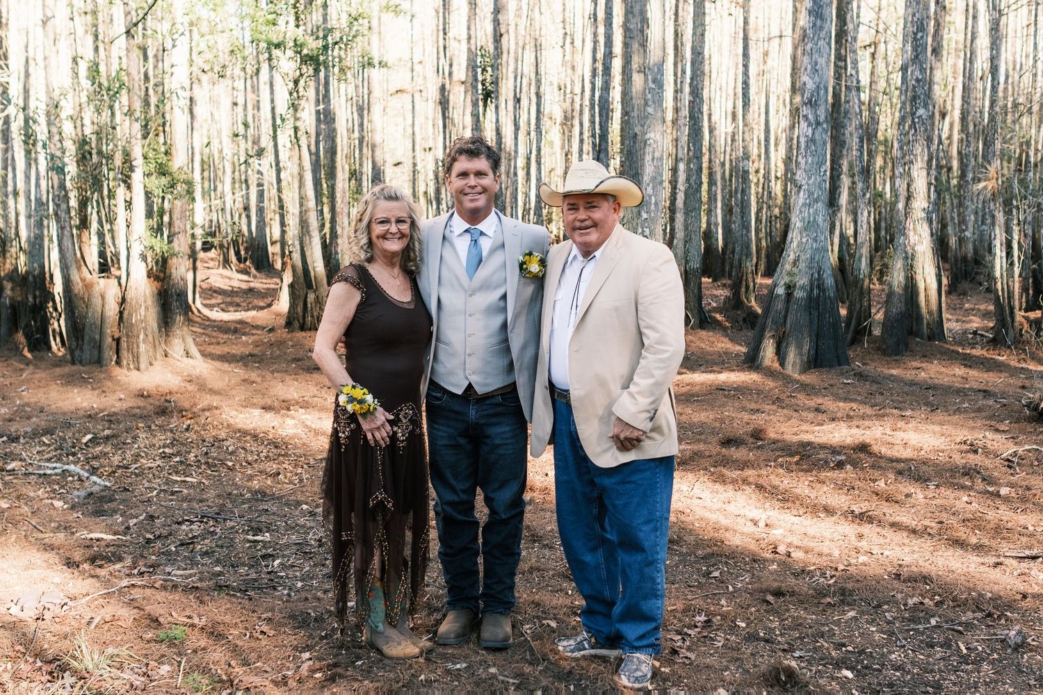 Groom in vest and tie poses with parents in a cypress tree forest at outdoor wedding.
