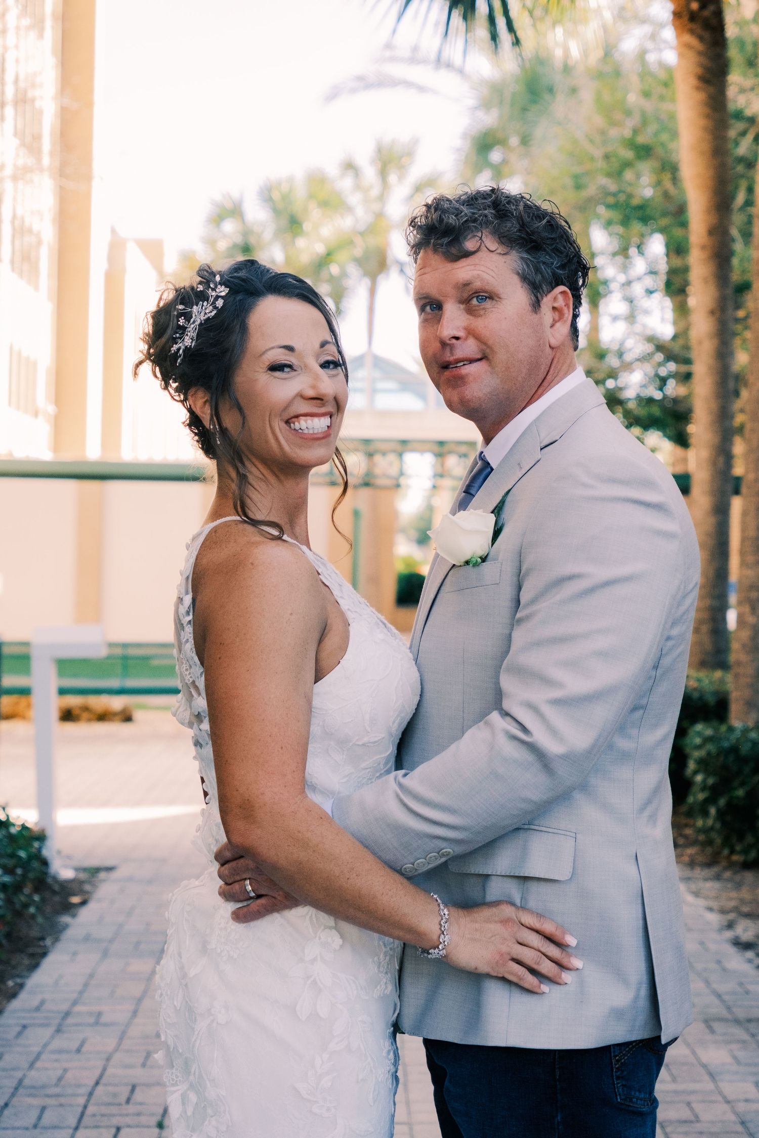 Smiling bride and groom embrace outdoors, she in lace gown with updo, he in tan suit with white boutonniere.